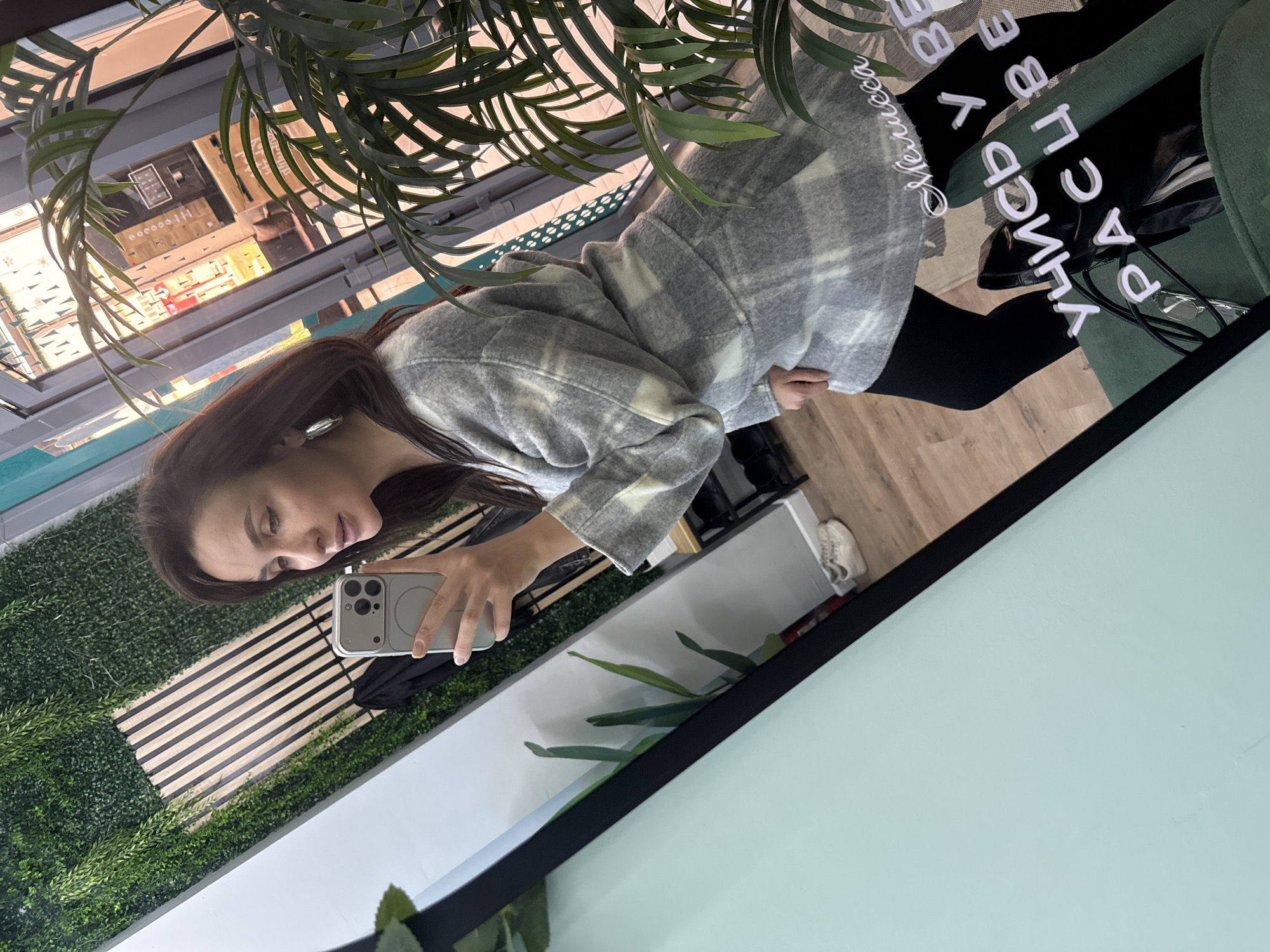 A woman is taking a selfie in a mirror. She is holding a white phone with a double lens camera. She is wearing a grey and white plaid jacket and black leggings. The mirror reflects a shop window and some greenery. In the mirror, backwards and at an angle, the text "Улица Раевского 3 Веселая Хамелесса" is visible. This likely indicates the image was taken on Raevskogo Street in Chaikovsky, Russia at the "Veselaya Khamelessa" store.