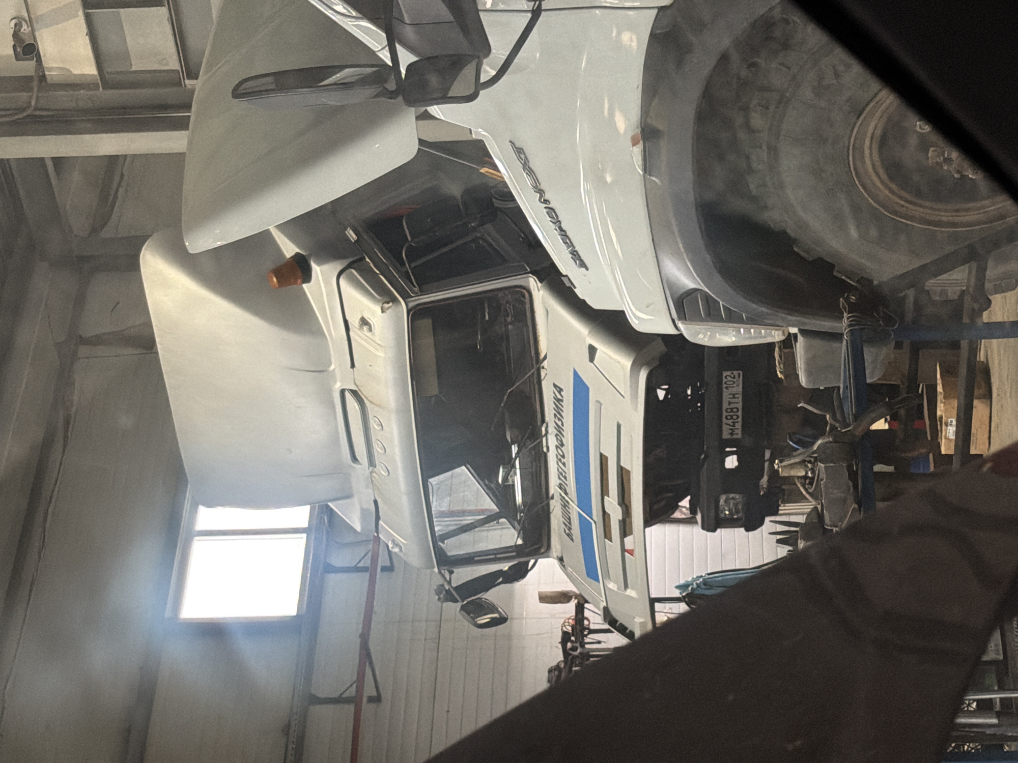 This is a close-up, vertically oriented, slightly blurry photograph of a white truck in a garage. The truck is elevated on a lift, with its underside visible. The front of the truck is to the left of the frame, and the rear is to the right. The cab of the truck is open, revealing some interior components. A large tire is visible at the top right of the image. The garage appears to be a workshop, with metal beams, conduits, and a window with bright light filtering through visible in the background. The overall lighting suggests it is daytime.
There is text visible on the truck. On the side of the cab, in Cyrillic, are the words "ВАШИР" followed by "ФИЗИКА". On the rear of the truck, a license plate is visible with the characters "M488TH 102".
The scene depicts a truck undergoing maintenance or repair in a garage setting. The focus is on the vehicle itself and its mechanical components. There are no people clearly visible in the frame. The environment suggests a professional or semi-professional repair facility. The image has a slightly dusty or hazy appearance, possibly from the garage environment or camera lens.