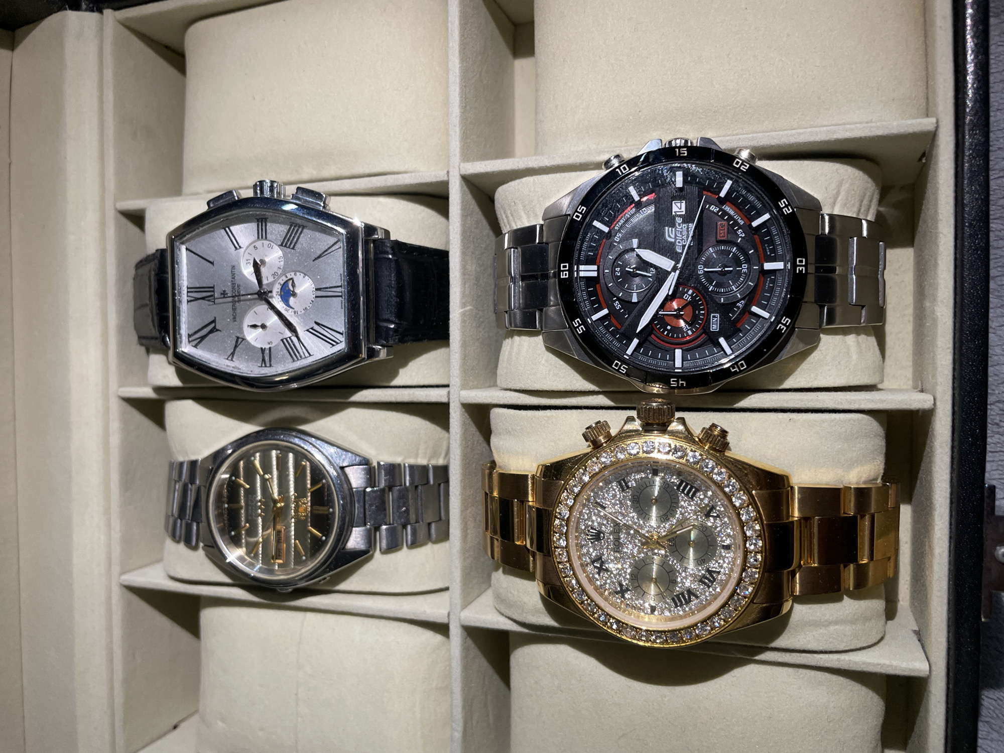 The image displays a collection of four wristwatches arranged in a storage case. The watches are positioned two per level, within individual compartments of the case. The compartment interiors are padded with a light-colored material.
The top-left watch has a silver-toned, rectangular case with a light-colored face, black leather strap and roman numerals. The top-right watch features a black dial, silver-toned stainless steel bracelet, and multiple sub-dials. The bottom-left watch has a silver-toned case and bracelet with a skeletonized dial revealing the watch's mechanism. The bottom-right watch is gold-toned with a gold bracelet and a dial adorned with crystals.
The image does not show any people or provide information about the location in Krasnoyarsk, Russia, or interactions.