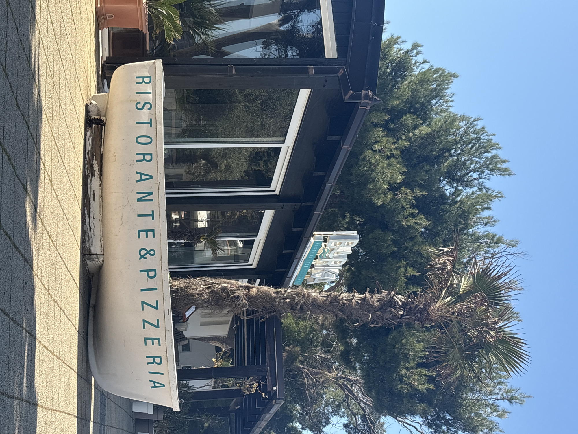 The image shows the exterior of a restaurant in Bar, Montenegro. A sign constructed from an old boat hull reads "RISTORANTE & PIZZERIA" in vertical, blue lettering. The restaurant has a modern design with dark framing and large windows. A dead or dying palm tree stands in front of the building. The sky is clear and blue. The building facade is light. The area around the restaurant is paved. The camera angle is looking up towards the restaurant.