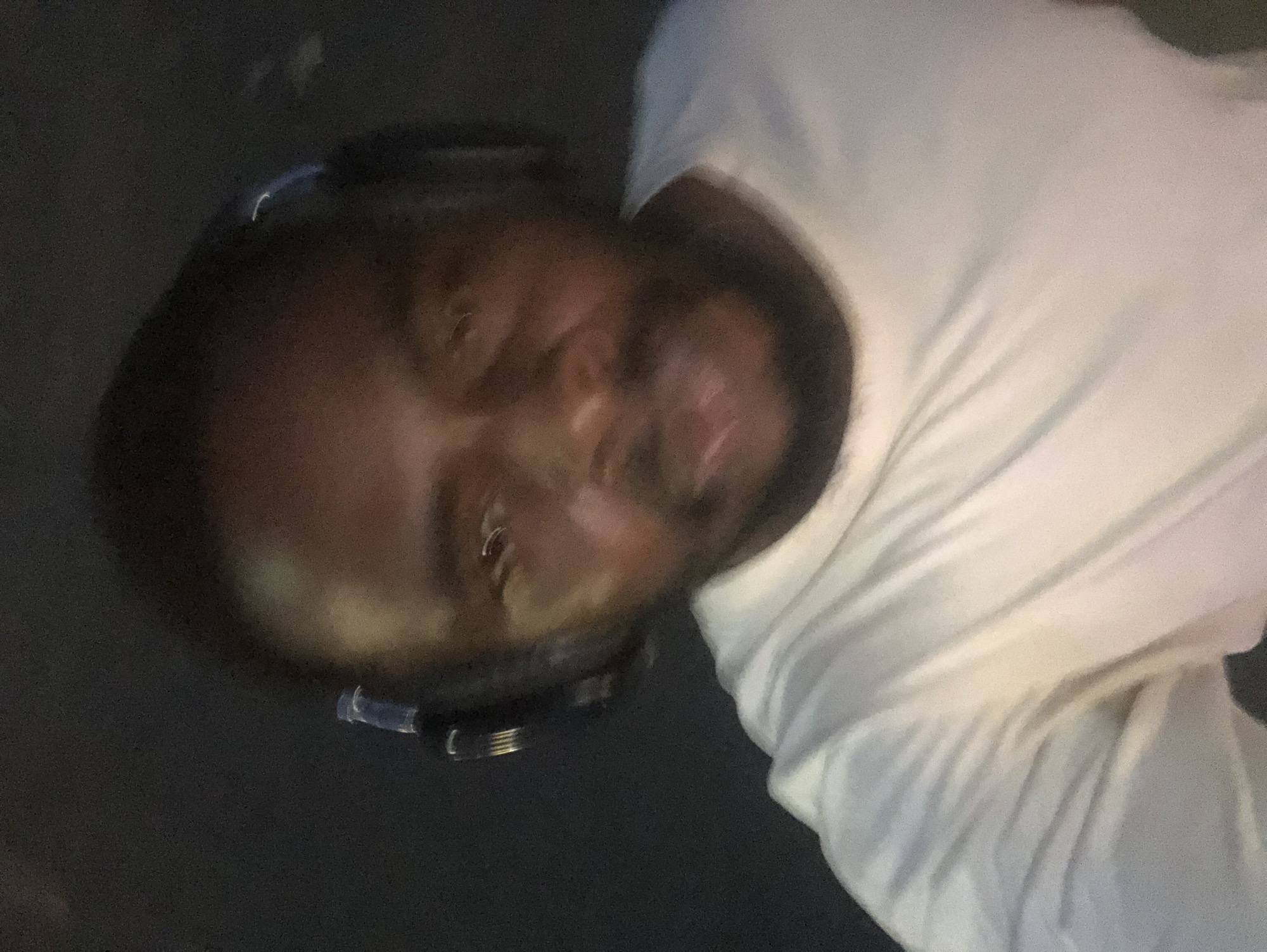 The media file displays a headshot of a person, likely male, wearing black over-ear headphones. The person has dark skin and is looking directly towards the camera with a neutral to slightly smiling expression. The face is somewhat blurry and soft-focused, making distinct features challenging to discern. They are wearing a light-colored, possibly white, long-sleeved shirt with visible folds and wrinkles around the collar and chest area.
The setting appears to be very dimly lit, with the background almost completely dark and devoid of details, suggesting low light conditions, possibly indoors at night or in a poorly lit room. The lighting casts some highlights on the person's face and the headphones, with a slight reflection visible on the headphone band. The overall image quality is dark, grainy, and slightly out of focus. The perspective is tilted, as if the person is reclining or the photo was taken at an angle.
The activity involves the individual wearing headphones, implying they are listening to audio. There is no visible text in the image. While the location context is given as Karu, Nigeria, there are no specific visual cues within the image itself to confirm this geographic location. The time of day appears to be evening or night due to the pervasive darkness.