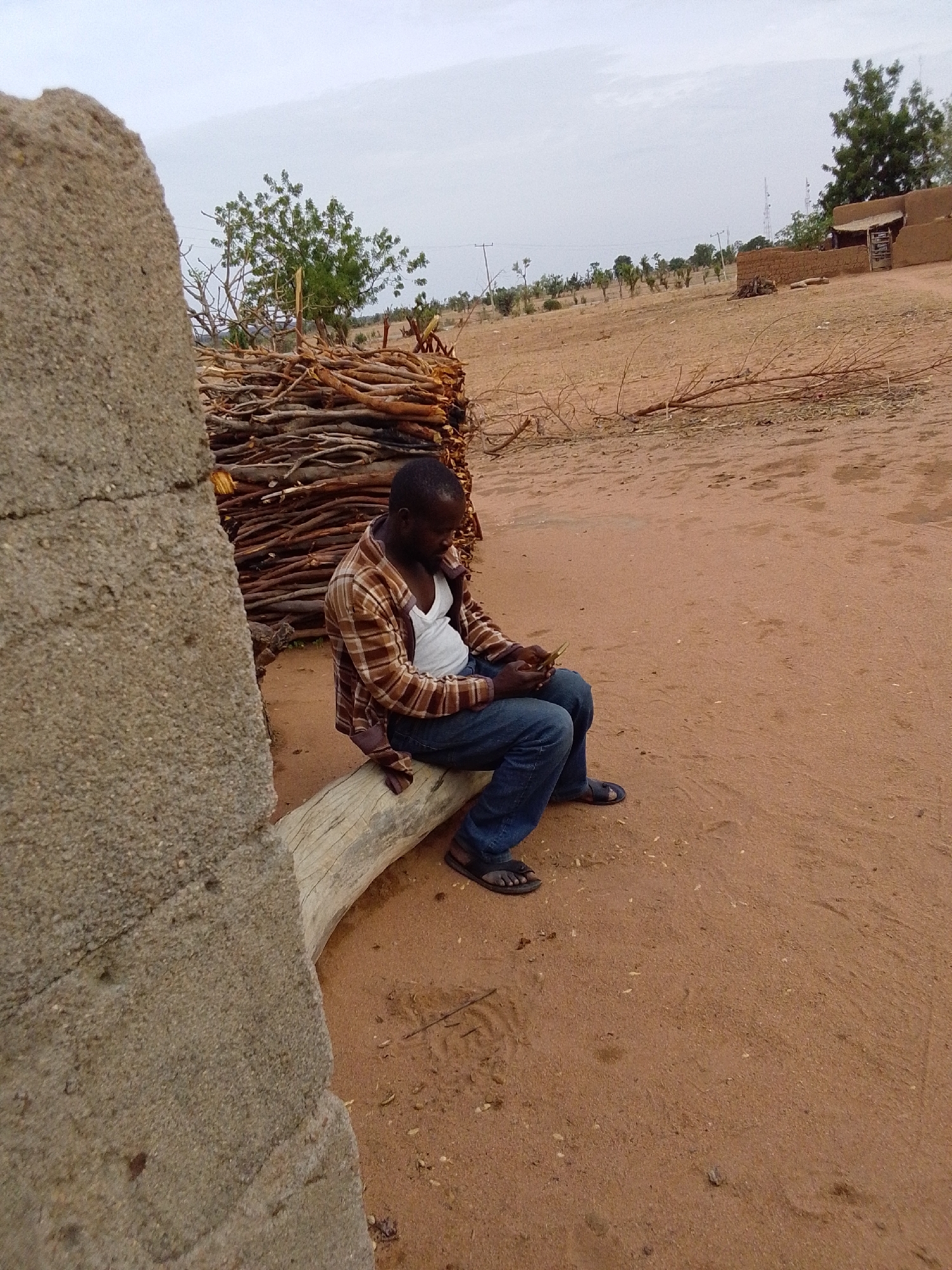 The image shows a man sitting on a log in what appears to be an outdoor rural setting in the city of Mulke Barikin Sayejo, Nigeria. He is wearing a white tank top, blue jeans, a brown and tan plaid shirt, and dark sandals. He is looking down at a mobile phone in his hands. The man is positioned to the right of a concrete structure and in front of a pile of branches and logs. The background reveals a sandy, arid environment with scattered trees, shrubs, and a distant building. The sky appears overcast. The ground is reddish-brown soil.
