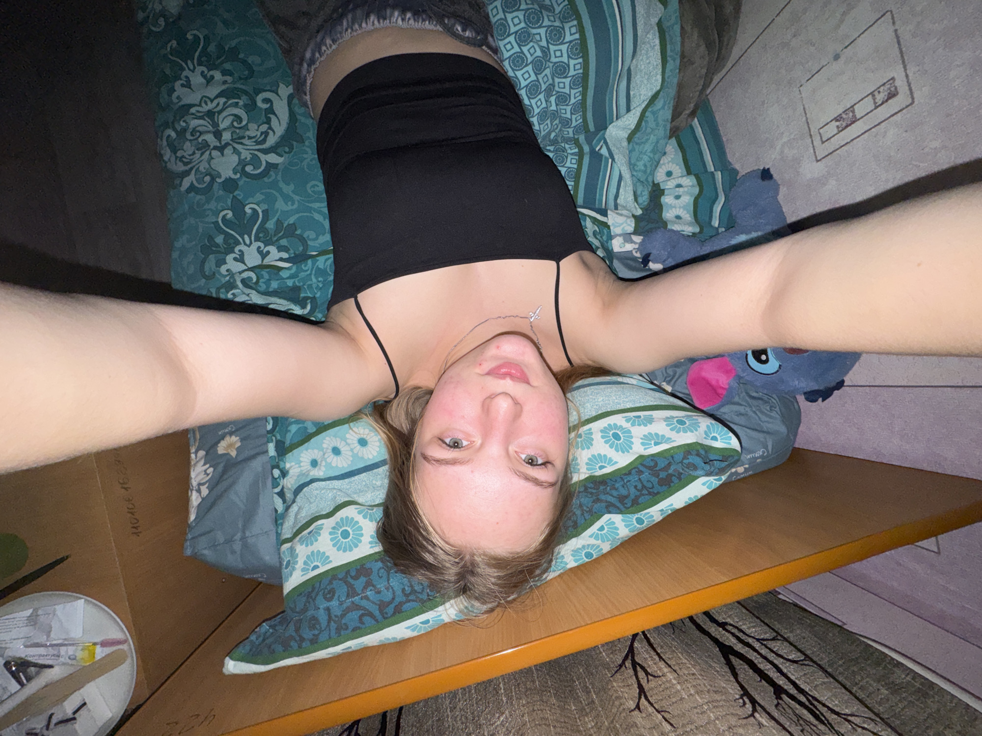 A woman with fair skin and blonde hair is lying on a patterned pillow on what appears to be a bed. She is wearing a black tank top and a silver necklace with a cross pendant. Her arms are extended towards the camera. The surrounding environment includes patterned bedding, a blue stuffed animal, a wooden surface with items on it, patterned floor covering, and a patterned wall. The location is Perm, Russia.