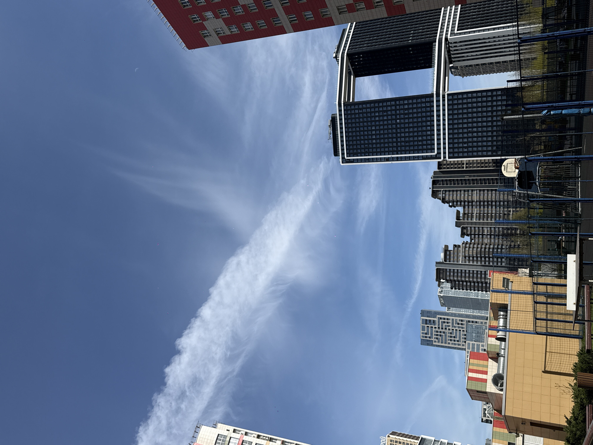 The image, taken in Перепечино, Russia, presents a dynamic upward view of a modern urban landscape under a bright, sunny sky. The sky is a clear blue, traversed by elongated, wispy white clouds or contrails. High in the sky, a faint crescent moon is visible.
Multiple contemporary buildings of varying heights and designs dominate the scene. On the left, a tall, red-colored building with numerous white-framed windows is partially visible. Towards the right, a prominent dark-colored skyscraper with distinctive white or light-colored structural outlines forms a large rectangular opening between two segments, creating a unique architectural silhouette. Further right and in the background, several other dark, multi-story buildings with glass facades and intricate window patterns, including one with a geometric design, rise towards the sky.
At a lower level, elements of an outdoor area are visible, including a basketball hoop and backboard, surrounded by blue and dark mesh fencing. Below these taller structures, lower-rise buildings feature more varied facades, such as one with horizontal stripes in beige, red, and green. Small green foliage is visible at the very bottom of the frame. Scattered small specks, possibly birds or distant objects, are present in the sky. Faint text "PÉO" is discernible on the edge of a building in the lower left. The overall impression is a bustling urban environment characterized by modern architecture under clear daytime conditions.