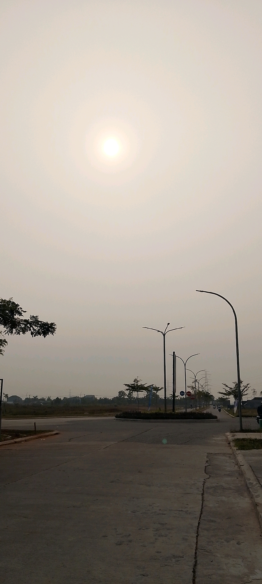 The image depicts an outdoor scene in the city of Satriamekar, Indonesia, characterized by an overcast sky with a bright sun visible in the upper center. The foreground shows a paved road, likely part of a new development, with a sidewalk on the right. Several modern streetlights are present, one tall, curved lamp on the far right and others with multiple arms in the middle ground. A circular median with vegetation is visible on the road. In the distance, there are residential buildings, trees, and power lines, suggesting an urban or suburban environment. The overall impression is of a developing area on a hazy or cloudy day.