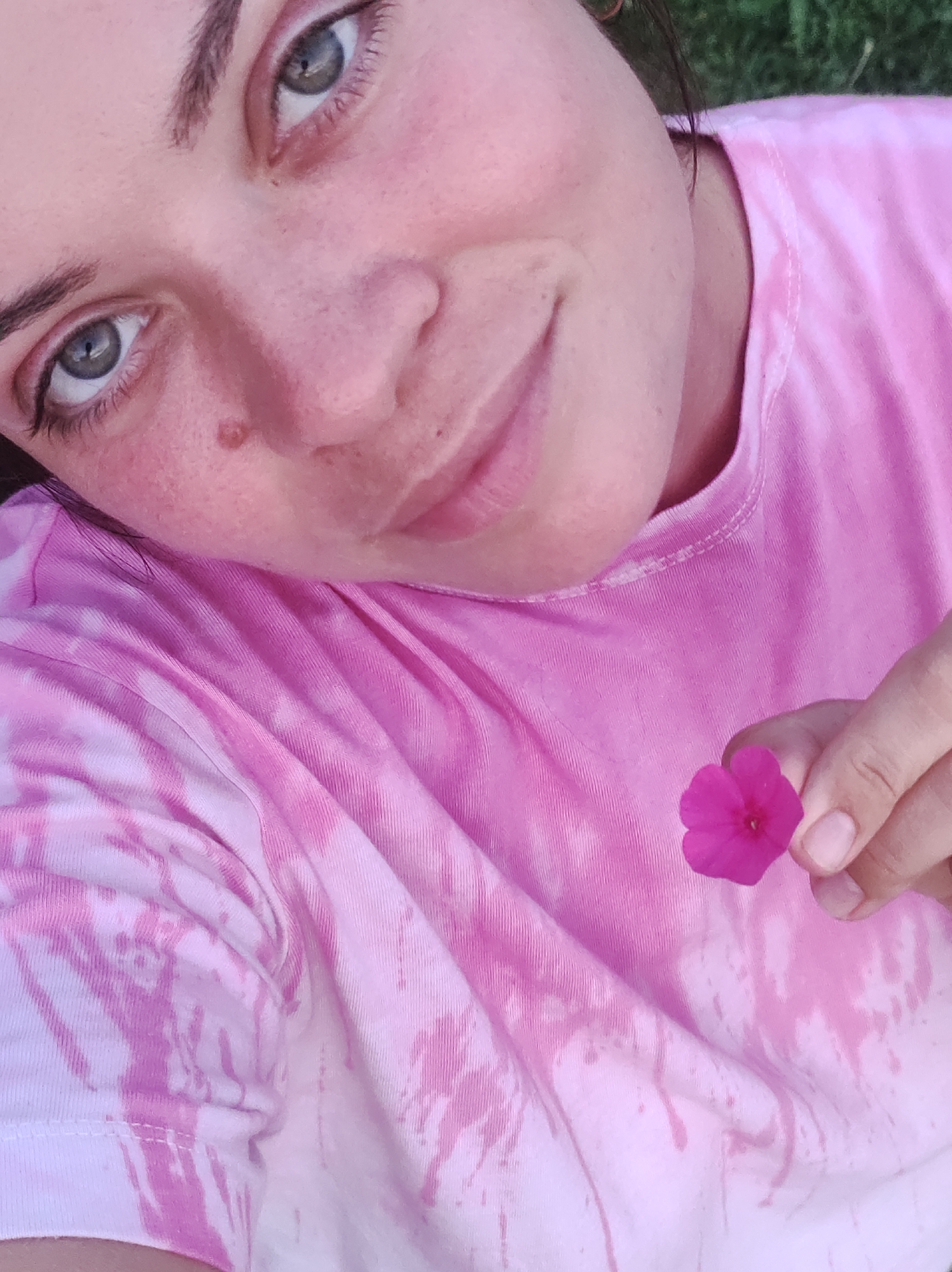 A close-up, high-angle shot shows a person's face and chest, wearing a pink tie-dye t-shirt. The person's left eye is looking directly at the camera with a gray iris and dark pupils. A small, brown mole is visible on the cheekbone. The person's right hand is holding a small, pink flower with four petals, with the thumb and index finger pinching the stem. The background is out of focus, showing green grass. The image was taken in the city of Fokino, Russia.