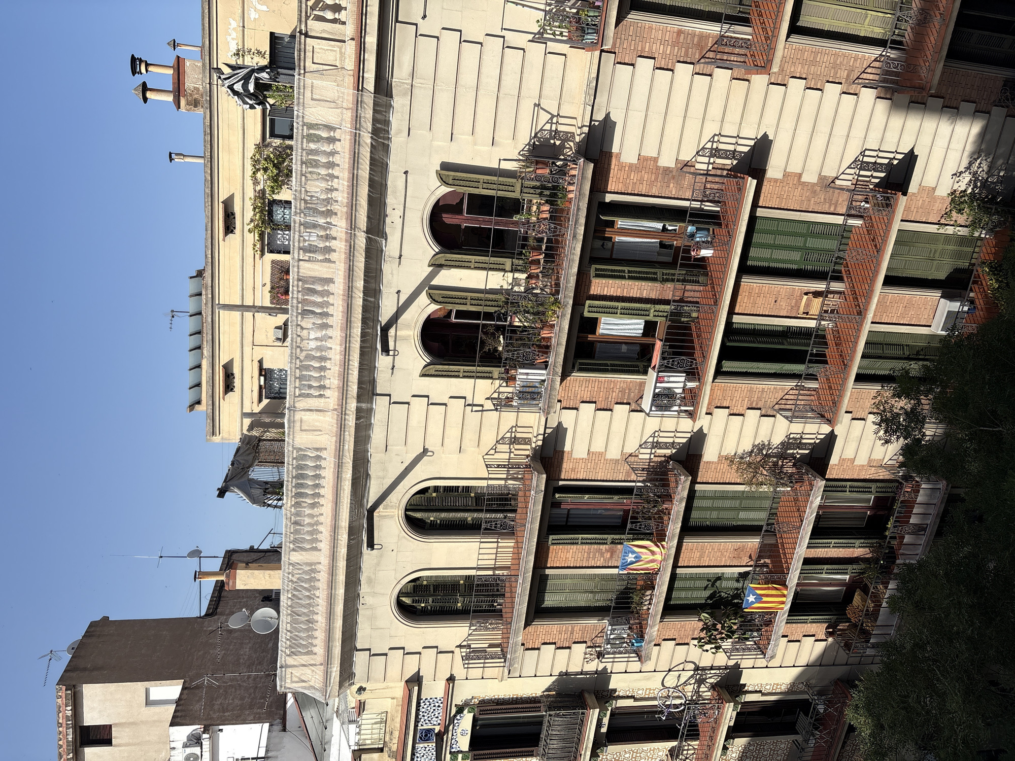 The image shows a multi-story building in Barcelona, Spain, viewed from an elevated perspective. The building features a light-colored facade with arched windows on the upper floors and brickwork on the lower levels. Several balconies, adorned with wrought-iron railings, are visible, some with potted plants and furniture. Two Catalan flags are displayed on balconies. The clear blue sky suggests a sunny day. The architectural style is typical of Barcelona, hinting at a historic district within the city.