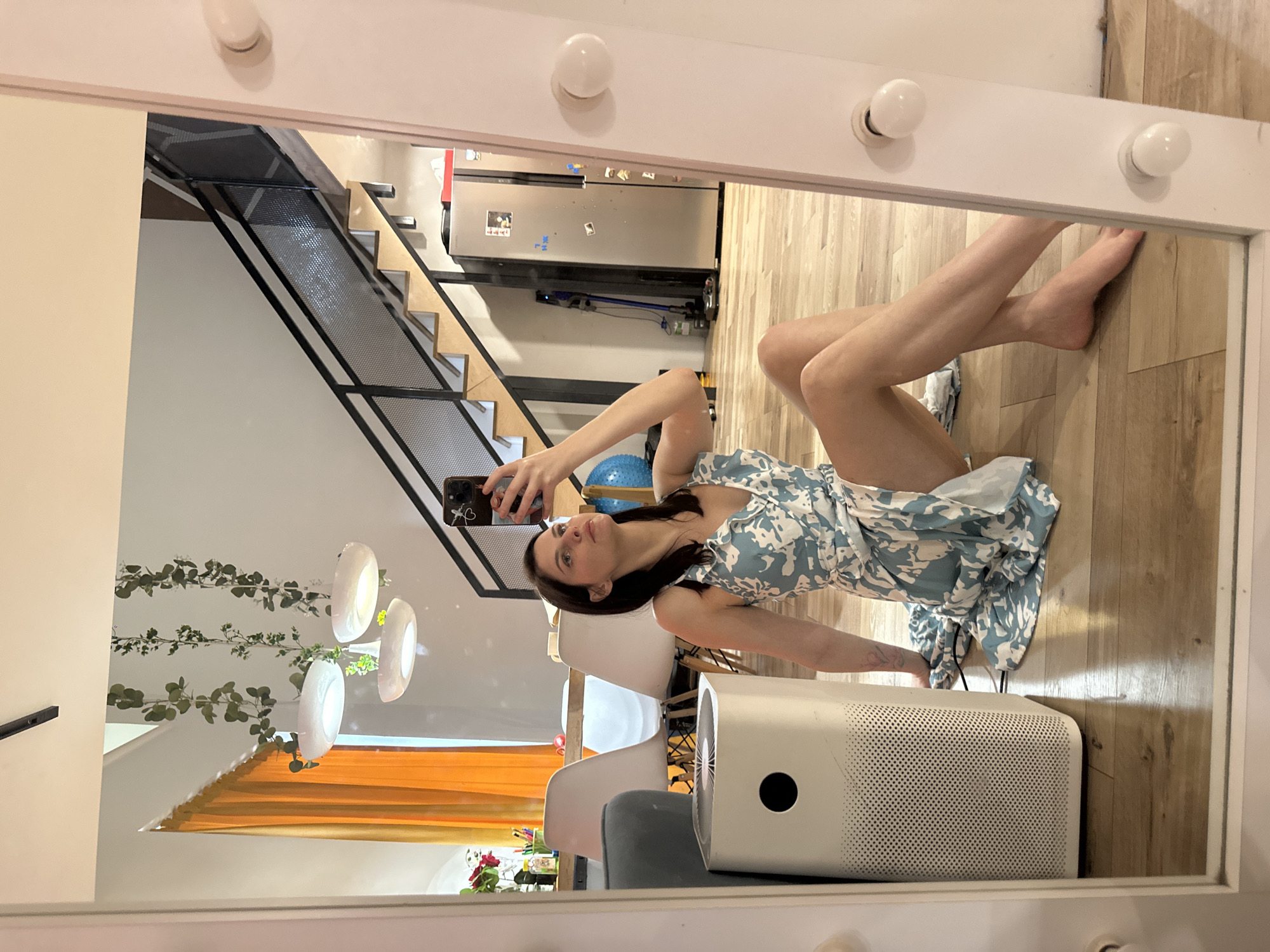 In a brightly lit room with wooden floors, a young woman is reflected in a large mirror. She is lying down, propped up on her arms, and wearing a blue and white floral dress. Her dark hair is shoulder-length, and she is looking towards the camera, holding a smartphone to take a selfie.
Behind her, also reflected in the mirror, is a modern interior. A staircase with black metal railings and wooden steps leads upwards. On the wall, a silver refrigerator is visible with some items attached to its surface. A Dyson vacuum cleaner is mounted on the wall beside it.
To the side of the woman, in the foreground of the mirror's reflection, is a white air purifier with a circular black vent. Beyond the air purifier, several white chairs are arranged around a table, and to the far left, a curtain with an orange hue is partially visible. Hanging from the ceiling are three large, round, white light fixtures. Green ivy plants are also seen trailing down a white wall.
The scene appears to be indoors, possibly in a home or studio, given the furniture and decor. The lighting suggests it could be daytime. The woman's pose and the selfie she is taking indicate a casual and self-aware moment.