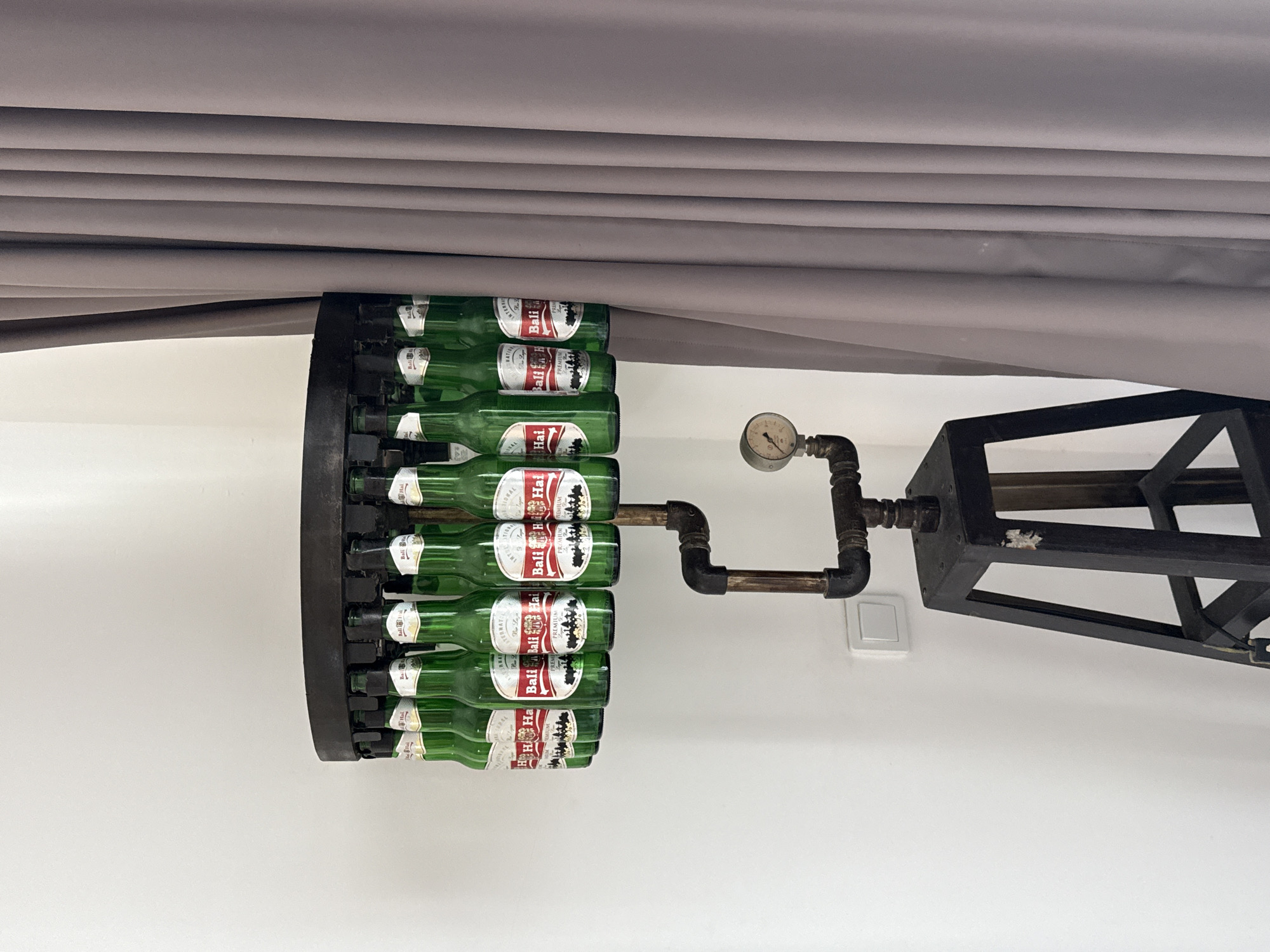 The image captures a creative decorative art piece attached to a white wall. The artwork comprises multiple green bottles of "Bali Hai Premium" beer arranged in a circular formation. These bottles are held within a black, circular framework. Extending from this circular arrangement is a metal pipe fitting, complete with a pressure gauge, which connects to a black, geometric metal structure that appears to be a decorative light fixture. A gray fabric hangs above this wall-mounted art piece. The location is the city of Unknown, Indonesia.