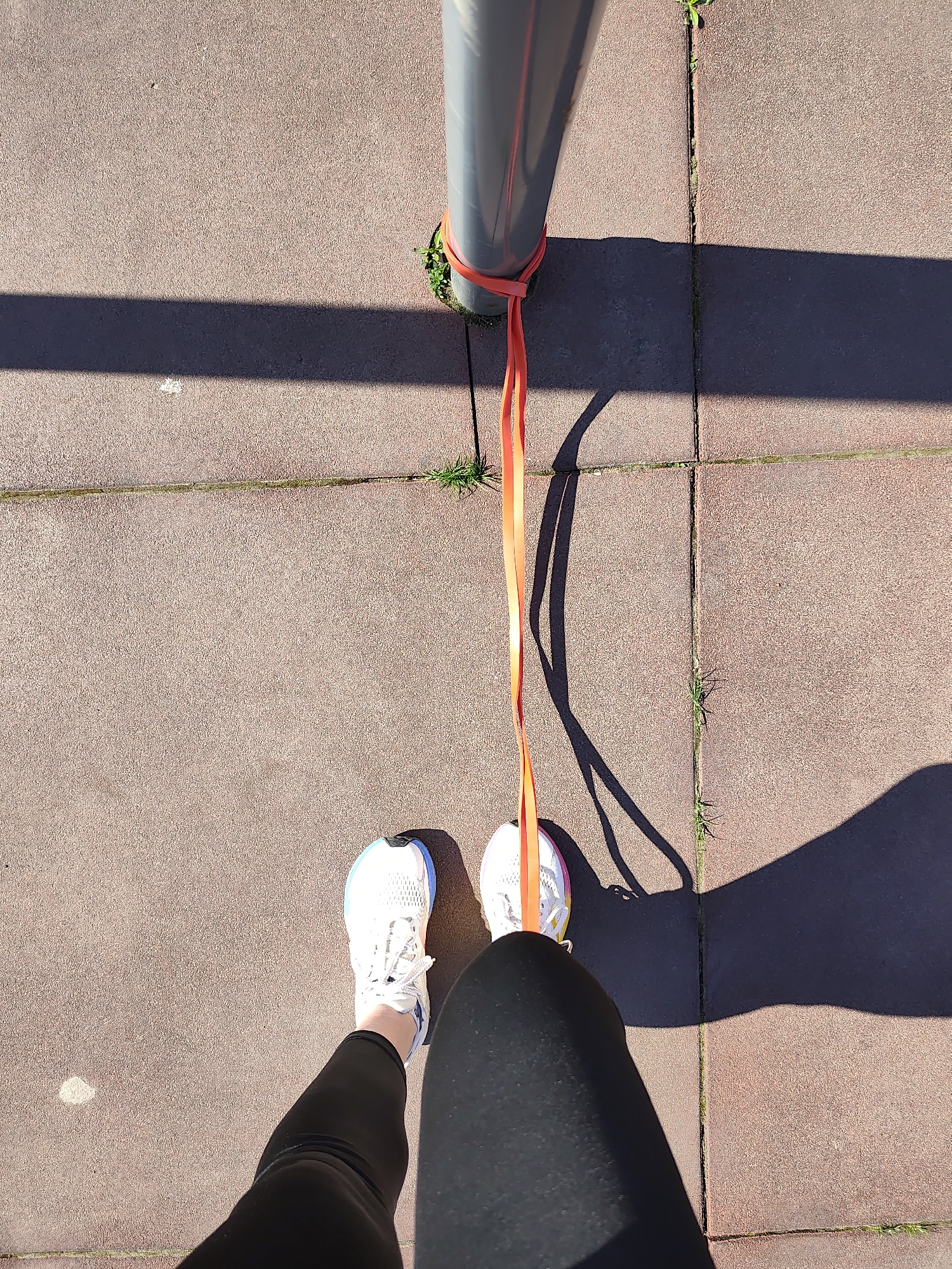 A first-person perspective photograph shows an individual standing on a paved surface, presumably performing a stretching exercise. An orange resistance band is looped around a grey metal pole and anchored to the person's left foot. The individual is wearing black leggings and white running shoes with blue and pink accents. The location is a sidewalk or similar paved area composed of square tiles. Sparse patches of grass grow in the cracks between the tiles. The shadow of the pole is visible, indicating sunlight. The city of Chaikovsky, Russia, cannot be independently confirmed from visual data.