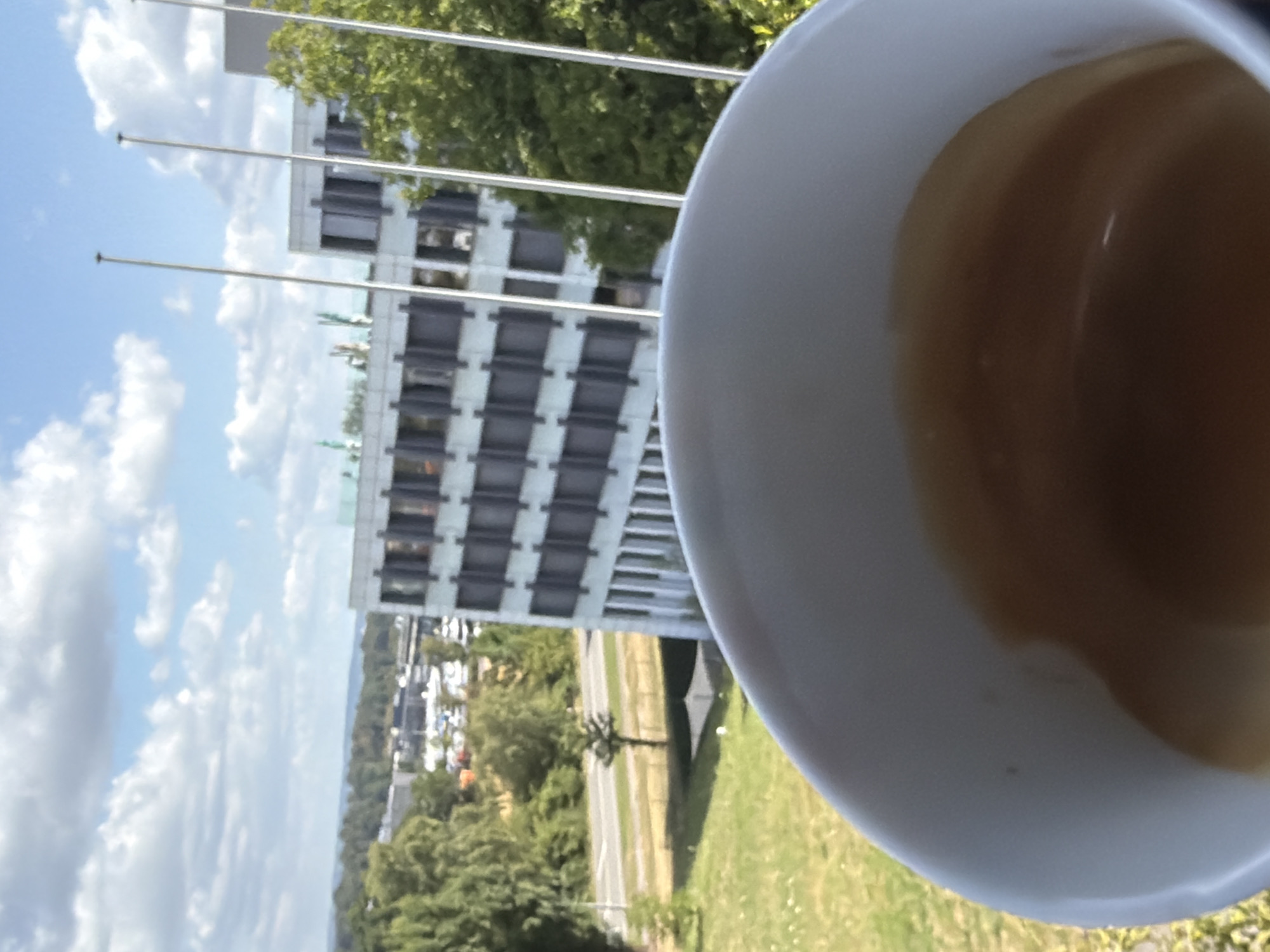 A white coffee cup with dark brown coffee is held in the foreground, obscuring a blurred view of a building and trees. The coffee is close to the rim of the cup and appears to have crema on top. In the background, a multi-story building with many windows is visible. Tall, thin poles stand in front of the building. Trees and a road with cars are also present in the midground. A green grassy area is in the foreground, suggesting an outdoor setting. The sky is blue with scattered white clouds. The image was taken in Leudelange, Luxembourg.