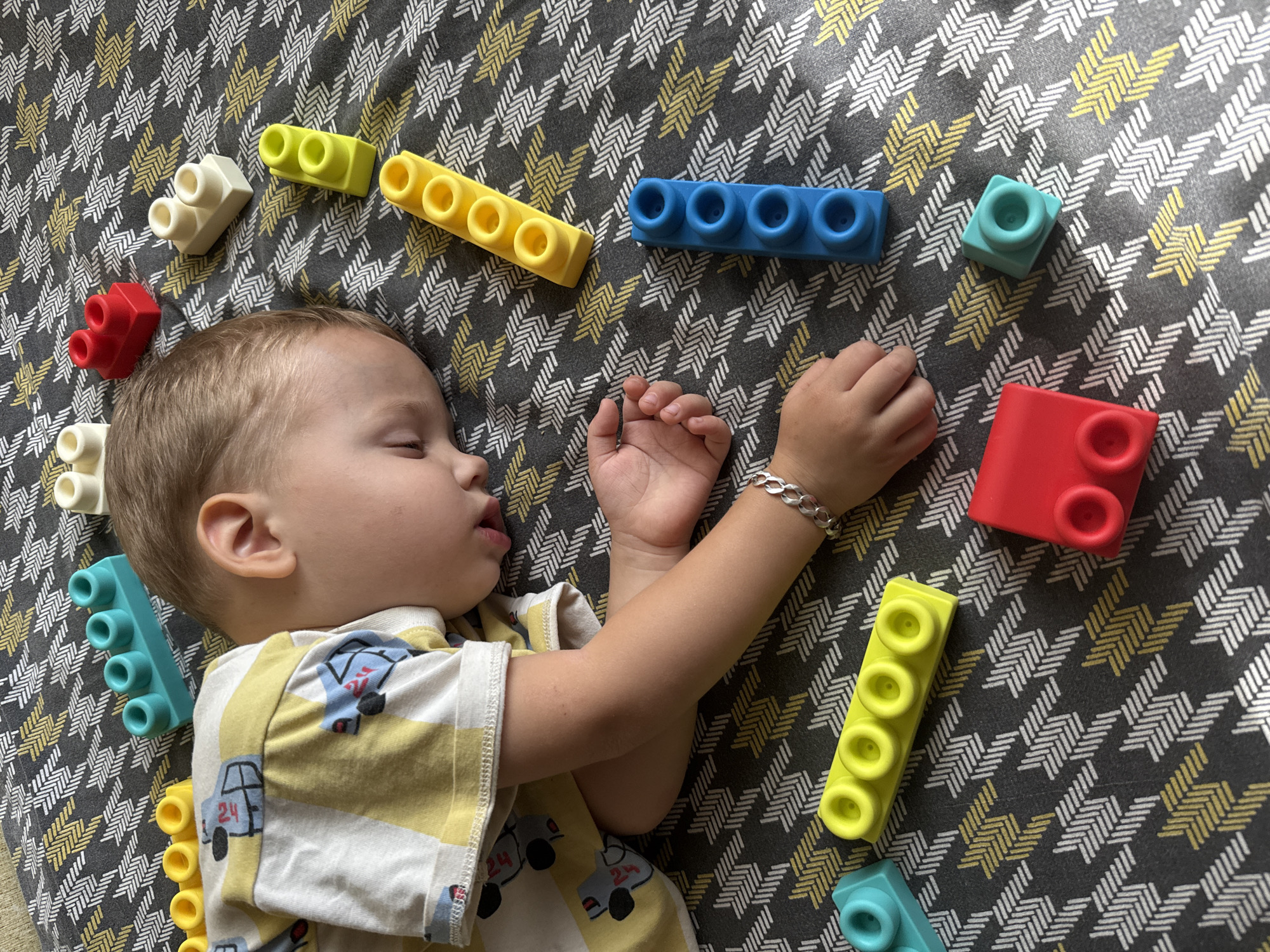 In Rusanovo, Russia, a young child sleeps on a patterned blanket surrounded by colorful building blocks. The child is lying on their side with their head resting on their arm. Their eyes are closed, and their mouth is slightly open. The child is wearing a yellow and white striped t-shirt with a cartoon car print. Building blocks of various colors, including red, yellow, blue, and white, are scattered around the child. The blanket has a grey background with a repeating geometric pattern of white and yellow zigzags. The lighting suggests it is daytime.