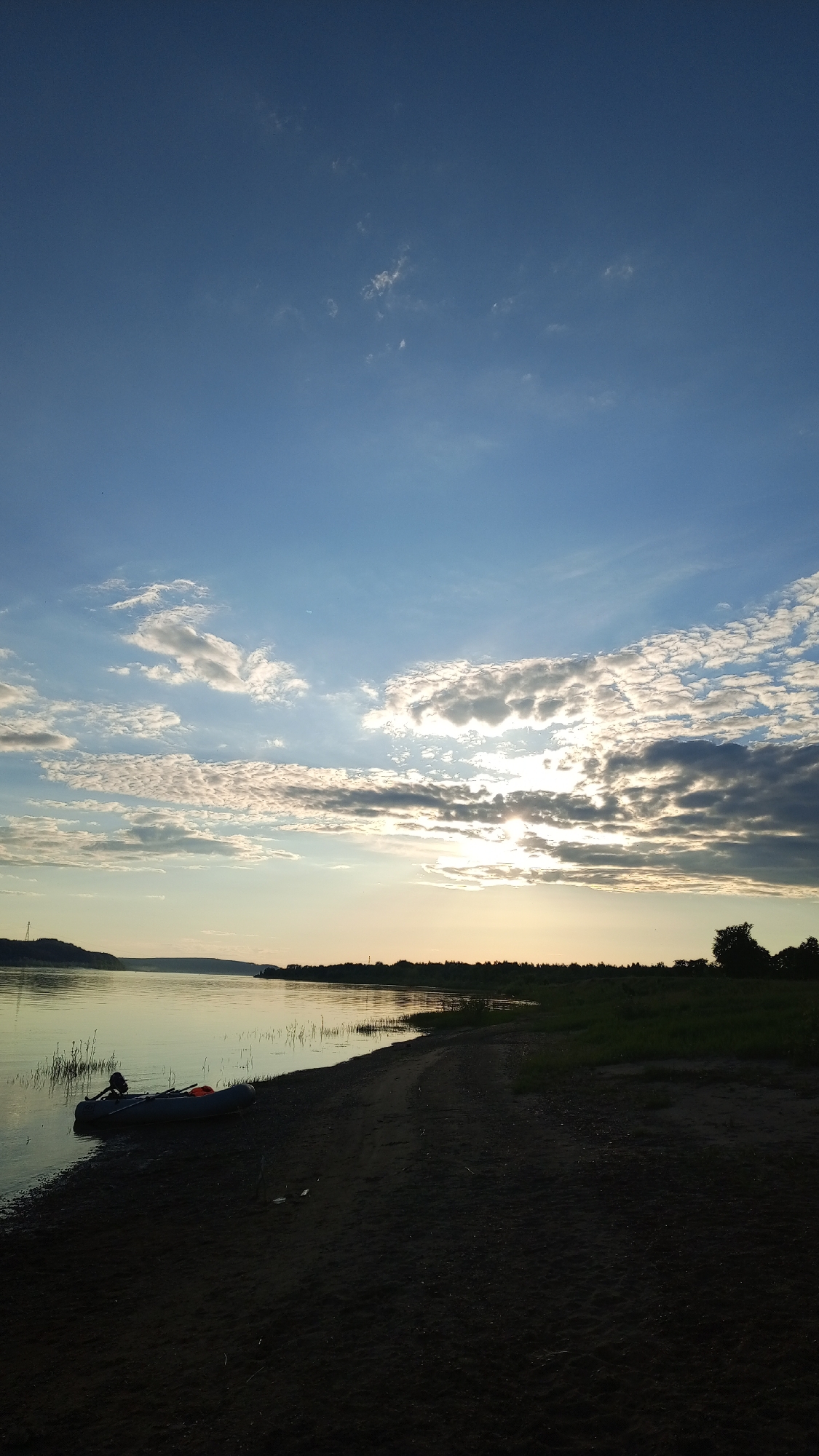 The image displays a tranquil waterside landscape at either sunrise or sunset, located in the vicinity of Шолья, Udmurtia, Russia, likely along the Kama River. The sky transitions from a deep blue at the top to lighter blue and warm golden hues near the horizon. The sun, low in the sky, is partially obscured by a formation of scattered cumulus and stratocumulus clouds, resulting in a bright, diffused glow. A wide body of water, reflecting the lighter parts of the sky, occupies the middle ground, showing subtle surface ripples. In the foreground, a dark sandy or dirt beach curves along the water's edge, transitioning to dark green vegetation on the right. A dark-colored inflatable dinghy, equipped with an orange cushion or seat and an oar or small motor, is partially pulled onto the sand at the water's edge on the left. Sparse reeds emerge from the water near the shoreline. The distant background features a dark, undulating landmass or hills silhouetted against the sky on the left, and a dense line of trees on the right bank. No human presence is observed in the scene.