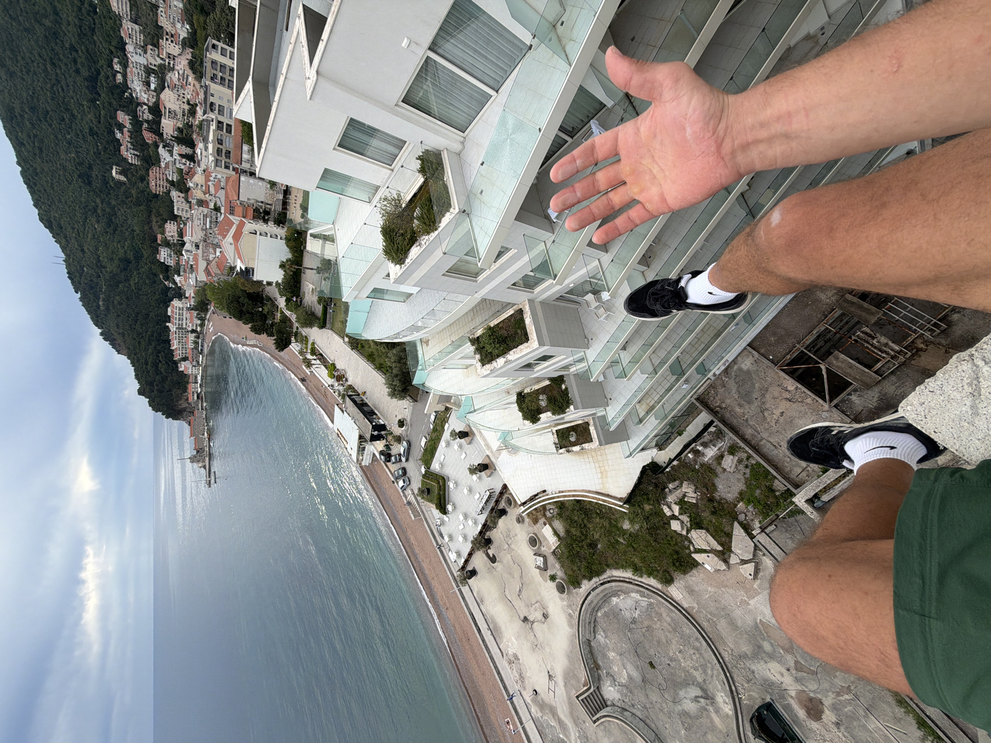 The image is a first-person view from a high vantage point, likely a building rooftop, in Petrovac na Moru, Montenegro. The scene encompasses a coastal area, a modern white building, and an individual.
Foreground: An arm and hand are extended, with legs and shoes visible, suggesting the person is on the edge. The shoes are black and white sneakers with white socks, and the person is wearing green shorts.
Environment: Below is a modern white building with glass balconies and greenery. A beach stretches along the coast, bordered by the Adriatic Sea. The water transitions from a lighter hue near the shore to a darker shade further out. A mountain covered in lush vegetation rises behind the town, with buildings scattered along its slope. The sky is overcast.
Location: Given the buildings and terrain, the location is Petrovac na Moru, Montenegro.