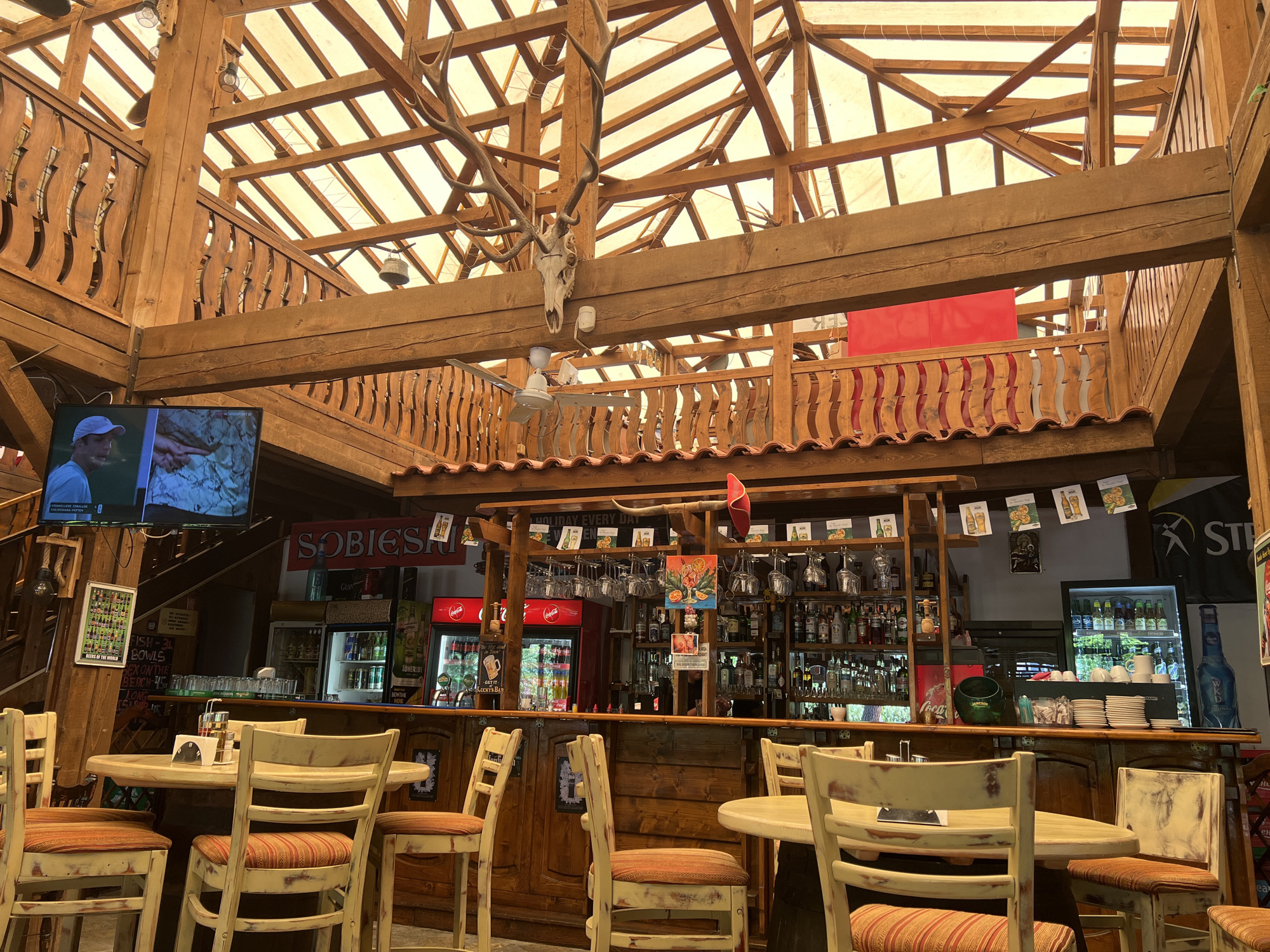 This detailed image captures the interior of a multi-level, rustic bar and restaurant, located in Nesebar, Bulgaria. The establishment is constructed primarily from heavy-set, light-brown finished wood, featuring prominent beams, support columns, and intricately carved wooden railings on a mezzanine level. The ceiling is composed of a framework of wooden beams supporting translucent panels, allowing abundant natural light to illuminate the space.
In the foreground and center, several light-colored, distressed wooden tables with matching chairs are arranged. The chairs feature striped fabric cushions in hues of orange, red, and cream. No individuals are present in the seating area.
The main bar counter, also crafted from dark-stained wood, stretches across the middle ground. Behind the bar, shelves are densely packed with various liquor bottles and an array of glassware, including hanging wine and cocktail glasses. Multiple commercial refrigerators displaying soft drinks (including Coca-Cola branded units) and other beverages are integrated into the bar structure. Draft beer taps are visible on the counter. A green bucket with "JAMESON" branding, a stack of white ceramic coffee cups, and other bar essentials are present. Signs for "SOBIESKI," "GUINNESS," "SOMERSBY," and "STELLA ARTOIS" are displayed around the bar area. Above the bar, a pair of animal horns and a red fabric decoration are mounted. A menu board lists "FISH - 3L BOWLS," "SEX ON THE BEACH - 6.50," and "LONG ISLAND..." cocktail prices. Another sign reads "HOLIDAY EVERY DAY."
Mounted high on a central wooden beam, a large, well-preserved deer skull with extensive antlers serves as a prominent decorative element. Directly below it, a white ceiling fan is suspended. To the left, a flat-screen television is mounted on a wooden column, displaying what appears to be a program featuring a man in a white cap on the left side of the screen and a close-up of a rock or textured terrain on the right. Below the television, a framed poster displays numerous miniature representations of various beers. The wooden railings on the upper level feature a distinctive wavy, carved design. The overall environment suggests a casual, lodge-like atmosphere.