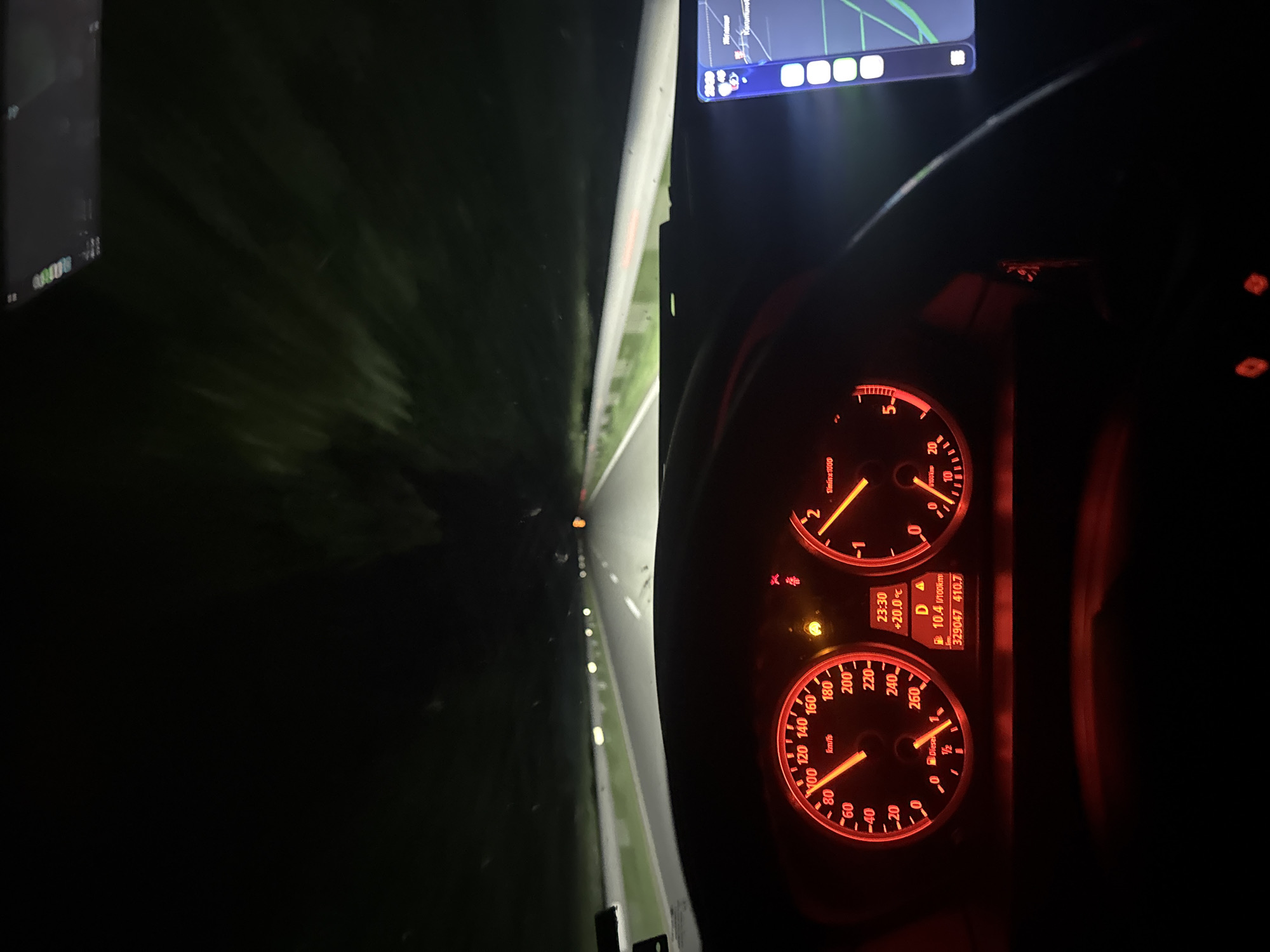 The image depicts the interior of a car at night, focusing on the dashboard and the view through the windshield. The dashboard instruments are illuminated in red, showing the speedometer reading approximately 100 km/h and the rev counter indicating a low engine speed. A digital display shows a fuel consumption of 10.4 l/100km and an outside temperature of +20.0°C. Through the windshield, a dark road stretches into the distance, illuminated by headlights. The location is likely a road in or around Stara Zagora, Bulgaria, based on the GPS navigation system visible in the upper part of the image.
