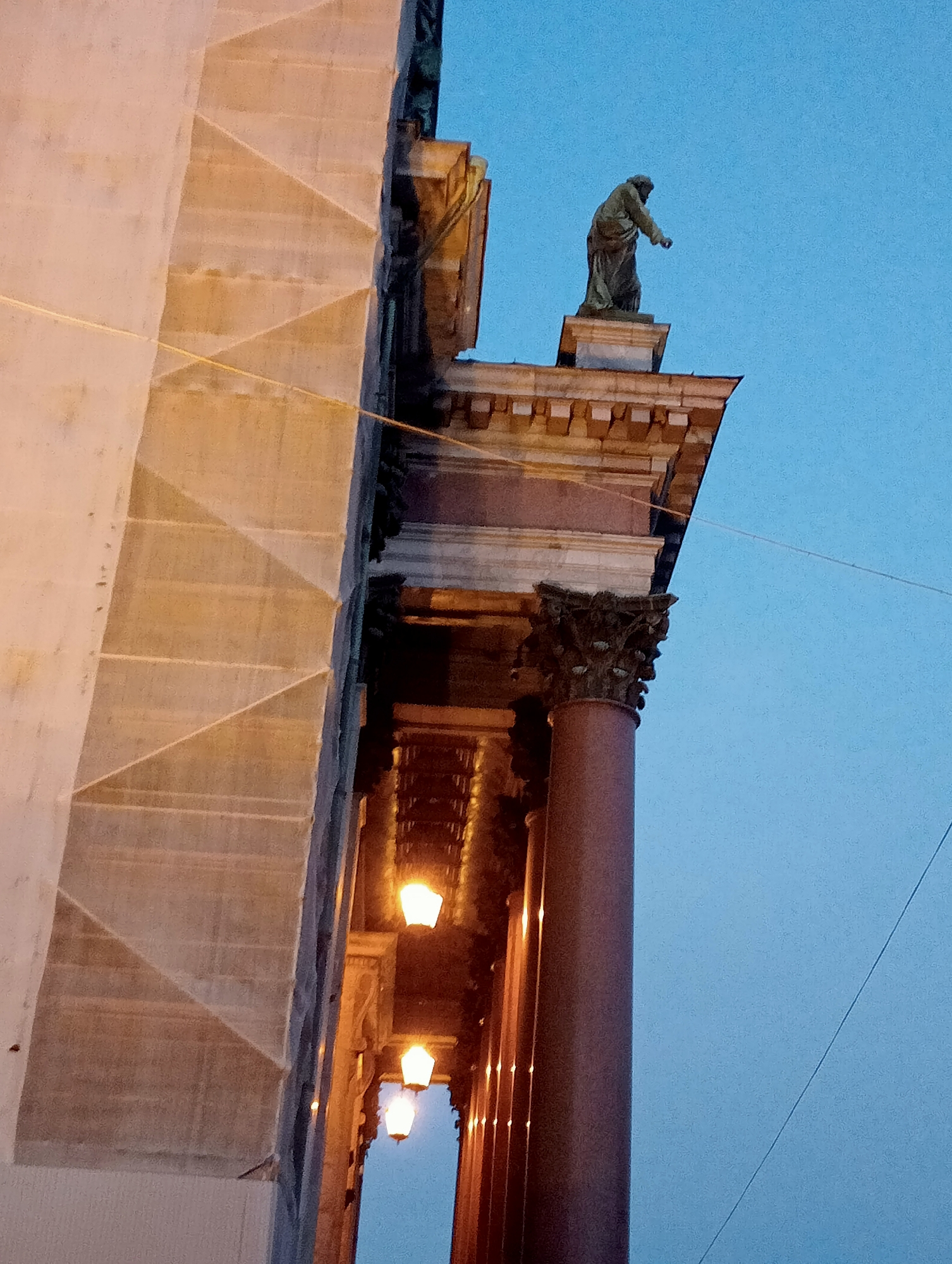 This is a photograph of the exterior of the Saint Isaac's Cathedral in Saint Petersburg, Russia, taken at dusk. The image shows a section of the cathedral's facade, including a large column and a statue of a figure standing on a ledge. The facade is made of stone and appears to be under repair, with scaffolding visible on the left side of the image. Several ornate streetlights are illuminating the lower sections of the building. The sky is a clear, pale blue, indicative of twilight. A thin wire runs diagonally across the sky in the upper right quadrant of the image.