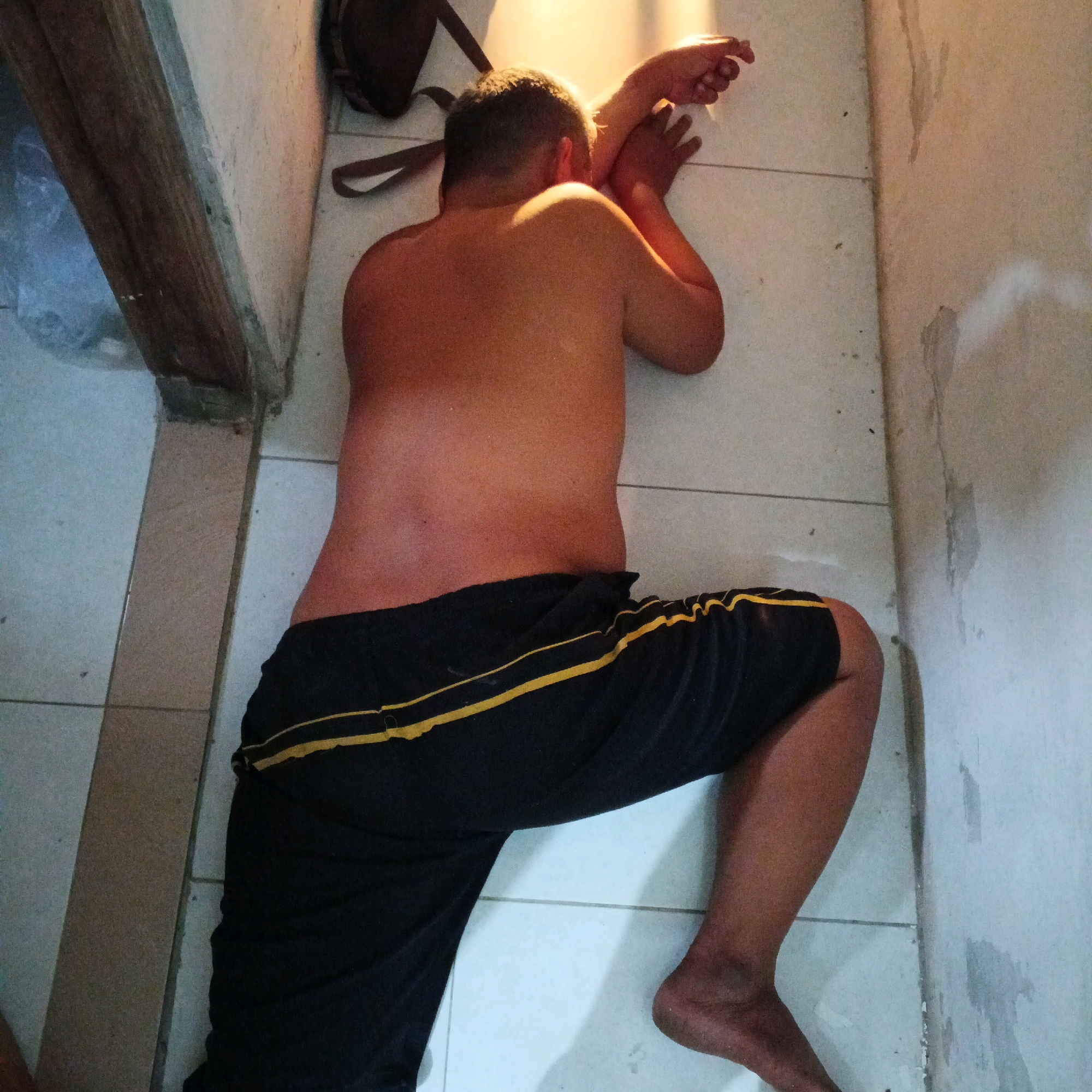 A shirtless male is lying on his side on a tiled floor in what appears to be an indoor space, possibly a room or hallway, with a light-colored wall and floor. The man is wearing black shorts with yellow stripes. Sunlight streams into the room from the right, illuminating the man's back and right arm. A dark bag is visible in the upper left corner of the image, resting against the wall. The overall lighting suggests it is daytime. The scene is located in Canggah, Indonesia.
