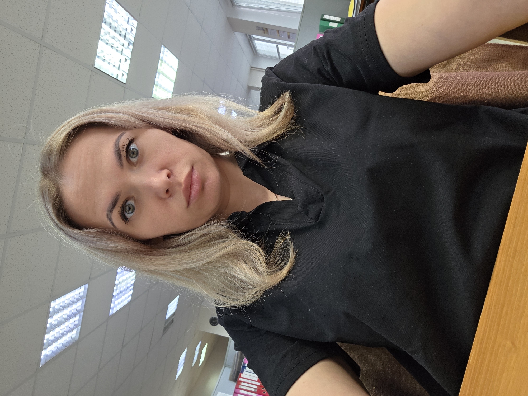 A woman with blonde hair, wearing a black shirt, is indoors. The background includes a ceiling with multiple square light panels and a desk. There is no specific information in the image to pinpoint the exact location within Chaikovsky, Russia.