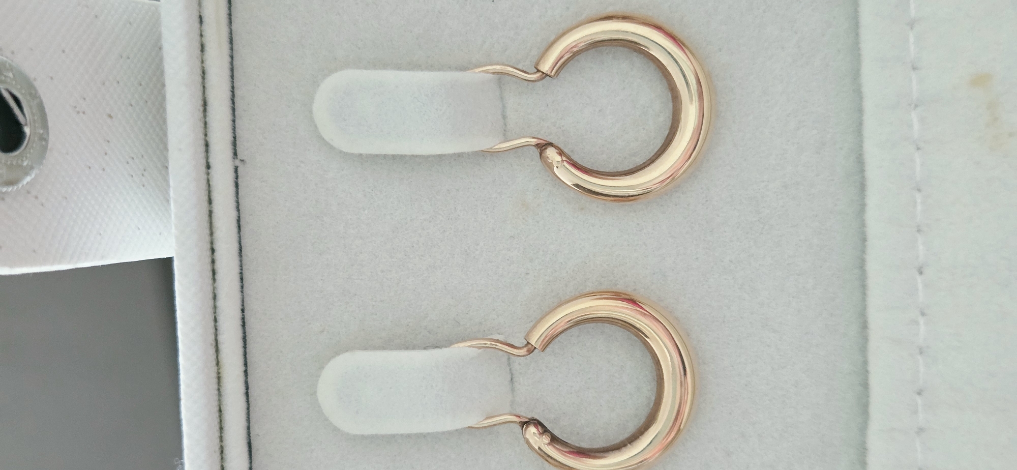 This image shows a pair of gold hoop earrings displayed in a white jewelry box. The earrings are gold-toned, with a rounded, tubular shape. They are attached to white, felt-like display cards. The background of the image is a light gray, textured surface, possibly the interior of the jewelry box. The lighting is even, highlighting the metallic sheen of the earrings. There are no people or animals visible in the image, and no specific location in the city of Lipetsk, Russia, can be identified from the visual information provided.