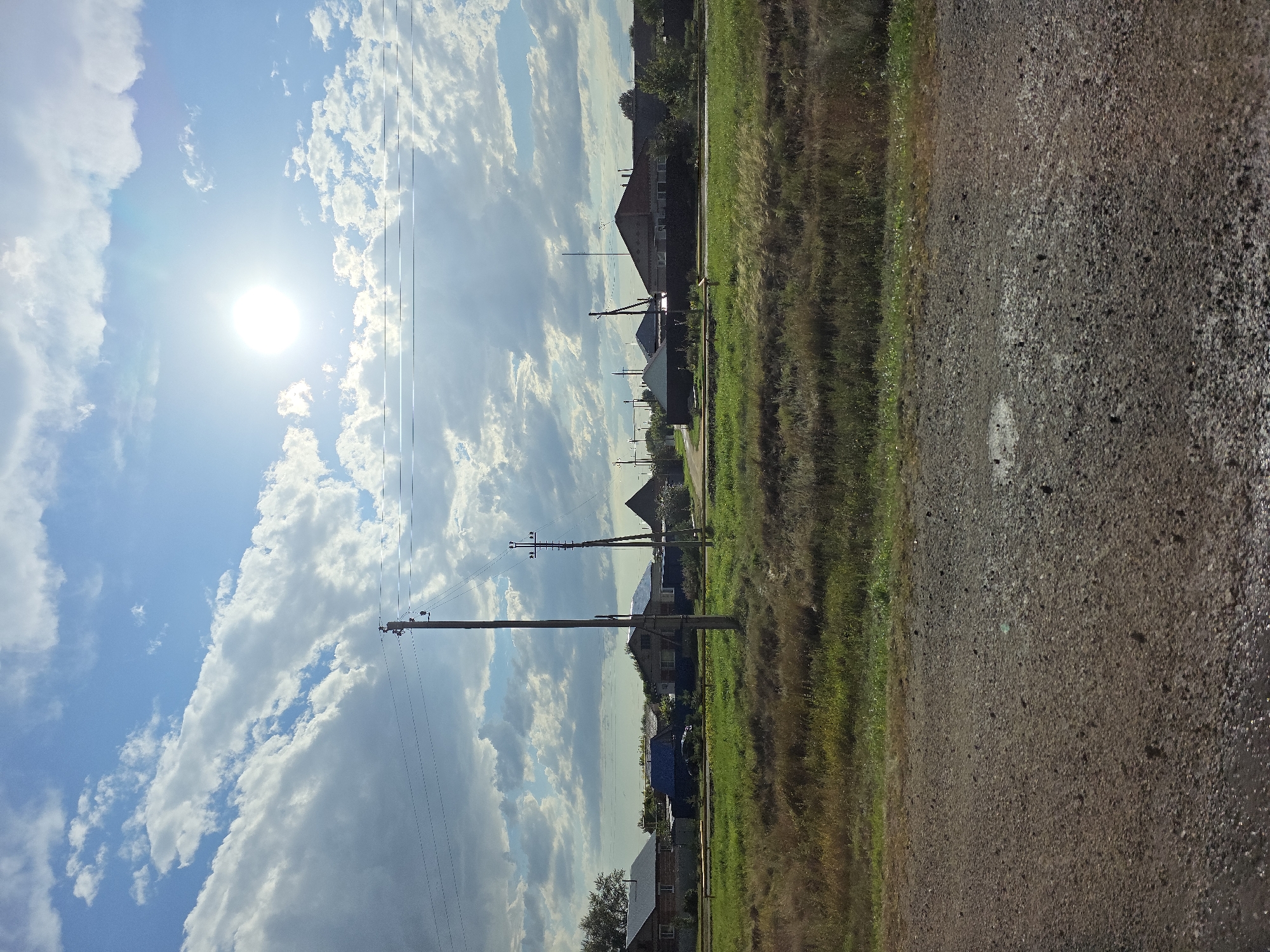 The image depicts a rural scene under a bright, sunny sky with scattered clouds. A dirt road occupies the foreground, with a grassy field beyond it. In the middle ground, a line of houses with pitched roofs are visible, interspersed with trees and utility poles supporting power lines. The sun is prominent in the upper right portion of the sky, casting a bright glare. The environment appears to be a residential area on the outskirts of a town or village, likely in Russia given the architectural style of the houses. The image is a still photograph taken from a slightly elevated perspective. No people or animals are visible in the frame.