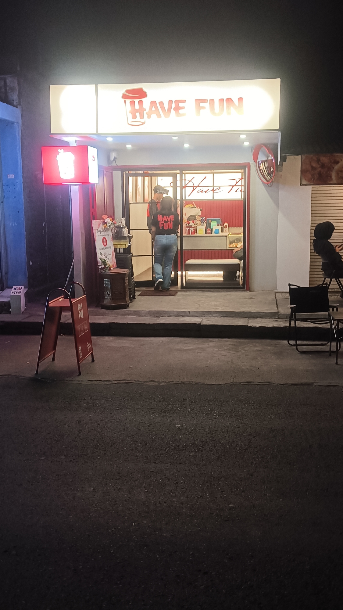 A bright, illuminated sign reading "HAVE FUN" with a stylized coffee cup logo is prominently displayed above the entrance of a shop. Below the sign, a red light-up box also features the coffee cup logo. The shop entrance appears to be a glass-fronted doorway with a person wearing a black t-shirt with "HAVE FUN" in red text and blue jeans walking inside. Another person, also wearing a "HAVE FUN" t-shirt, is visible behind the counter. Inside the shop, there are shelves stocked with various items and a red interior theme. Outside the shop, a red A-frame sign with "HAVE FUN" printed on it stands on the sidewalk. The scene is set at night, on a paved street in front of the shop, with a few outdoor chairs visible to the right. The overall impression is that of a beverage or snack shop. This scene is located in Sragen, Indonesia.
