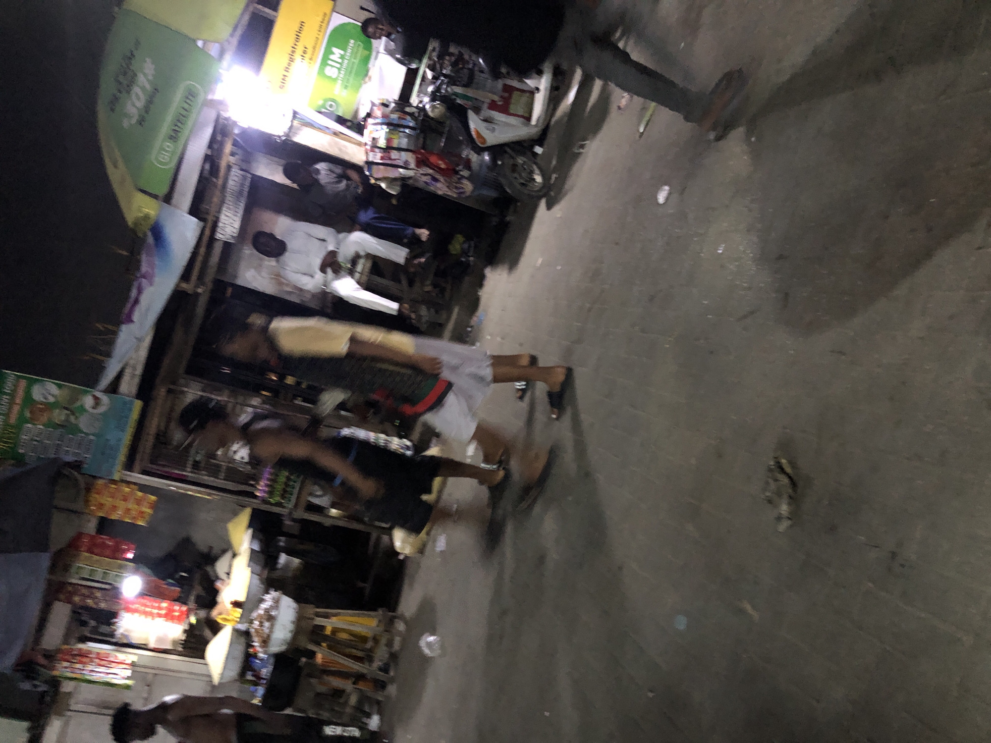 The image shows a street scene in Moba, Nigeria. Multiple pedestrians are present, engaged in walking and standing. Commercial activity is apparent, as evidenced by shops, displays of goods, and advertising signs. A parked motorbike is also visible. The overall environment suggests an active, urban area with pedestrian and vehicular traffic.