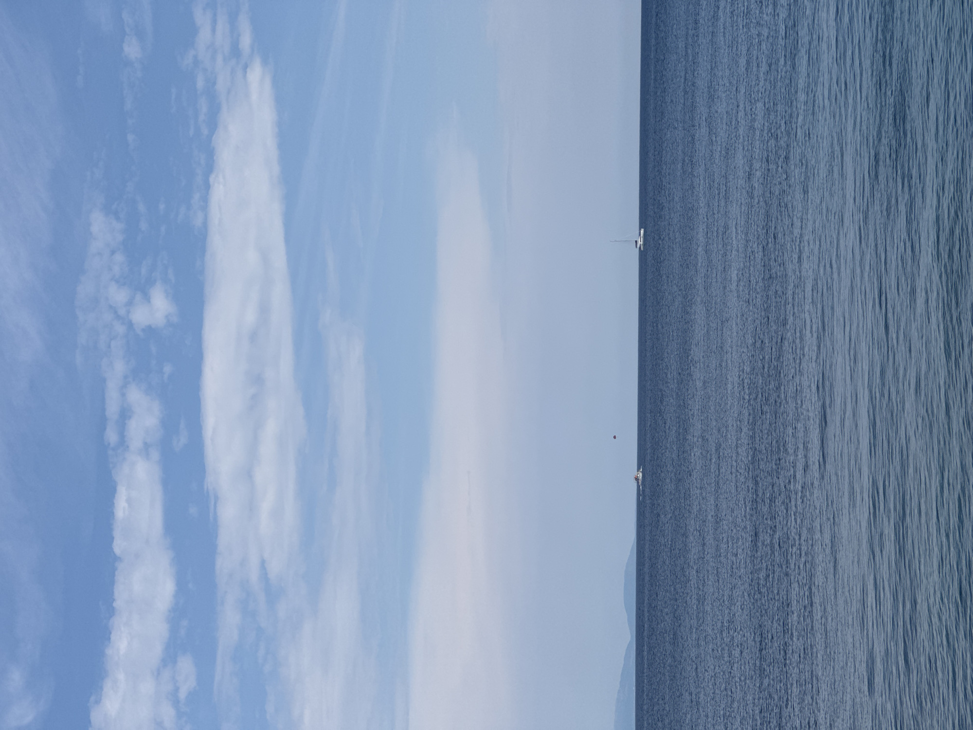 An aerial view shows a calm, deep blue sea under a bright sky dotted with white clouds. In the distance, a thin strip of land with rolling hills is visible along the horizon. Two boats are present on the water. One is a small white sailboat with a tall mast, positioned on the right side of the frame. To the left of the sailboat, a motorboat is visible, towing a small red parachute that is airborne, suggesting parasailing activity. The overall scene depicts a serene coastal environment. The location is identified as Dubraljevina, Montenegro.