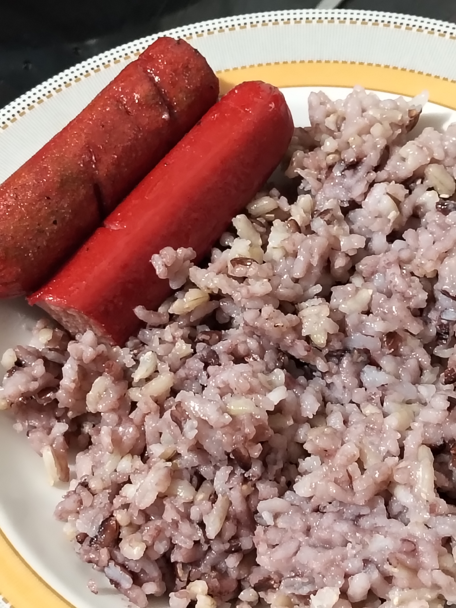A close-up, overhead shot of a plate of food shows two red hot dogs resting on a bed of purple rice on a white plate with a decorative yellow and gold rim. The hot dogs are slightly charred and appear to be split down the middle, revealing a lighter interior. The purple rice is glistening and has a textured appearance, with some grains appearing darker than others. The background is dark and out of focus. The image is likely taken indoors, possibly in a kitchen or dining area, and does not provide any specific visual cues to pinpoint a location within Davao City, Philippines.