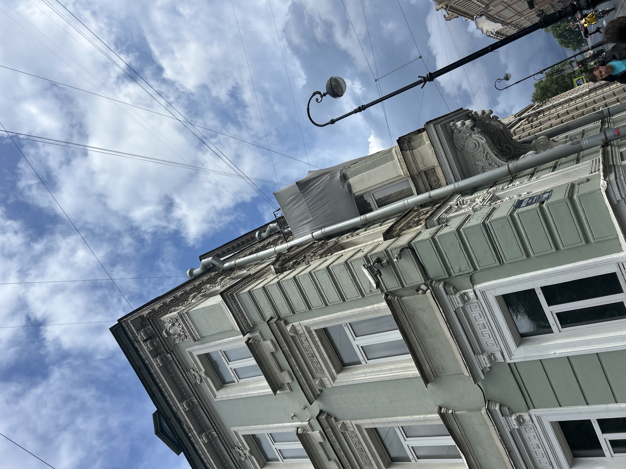 A low-angle, wide shot captures the side of a pale green building in a city. The building is ornate with white trim around windows and decorative moldings. Several windows are visible, many with white frames and panes reflecting the cloudy sky. A downspout runs vertically along the side of the building. Above the building, a tangle of overhead wires crisscrosses the cloudy blue sky. A black street lamp with a rounded glass shade stands to the right, its curved pole topped with a decorative finial. In the background, a street is visible with a traffic light, trees, and the facades of other buildings. A few people are present, including a man wearing a blue shirt and a woman in a yellow jacket on a bicycle. The overall impression is of an urban streetscape under a dramatic sky.
