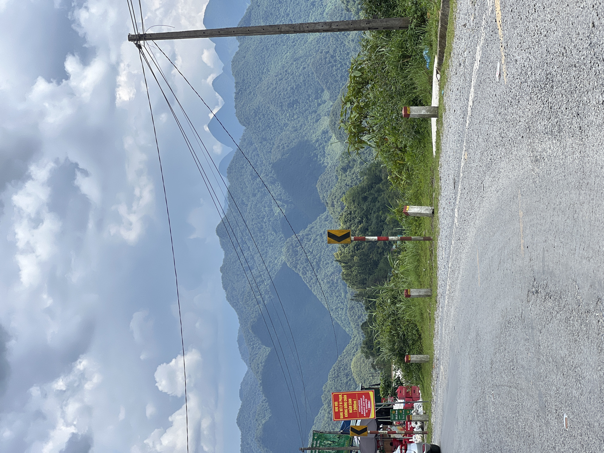 The image shows a scenic mountain landscape in Quản Bạ Commune, Vietnam, with lush green peaks under a cloudy sky. A paved road curves through the foreground, with a gravel shoulder and several small concrete posts along its edge. In the middle ground, there are signs, including a red one with Vietnamese text, a yellow directional sign, and some sort of market stall or roadside vendor with stacked crates. A utility pole with multiple wires stretches across the frame. The background is dominated by layered, verdant mountains that recede into the distance.