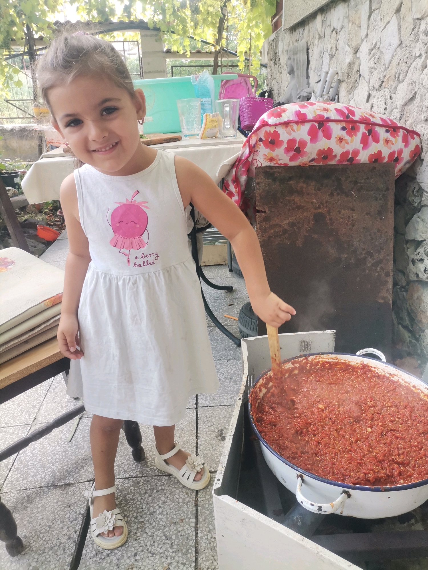 In Obrochishte, Bulgaria, a young girl with brown hair pulled back stands in front of a large pot filled with a red, chunky mixture, possibly food being cooked outdoors. She wears a white sleeveless dress with a pink strawberry graphic and white sandals with floral embellishments. Her right arm is extended, holding a wooden spoon, and she appears to be stirring the contents of the pot. The background reveals a stone wall, greenery from overhead vines, and various items on a table, including glasses and a basket. The scene suggests a domestic setting, possibly a backyard or outdoor kitchen area.