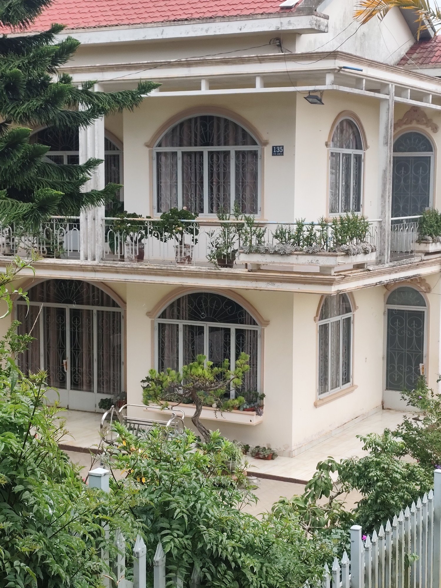 A three-story building with a red tiled roof and cream-colored facade is shown. The building has multiple arched windows with decorative metal grilles. Balconies with white railings and potted plants are visible on the upper floors. A number "135" is displayed on a small plaque near one of the windows. Lush green trees and foliage surround the building, and a white picket fence runs along the bottom of the frame. The scene appears to be a residential property in Lang Biang - Đà Lạt, Vietnam.