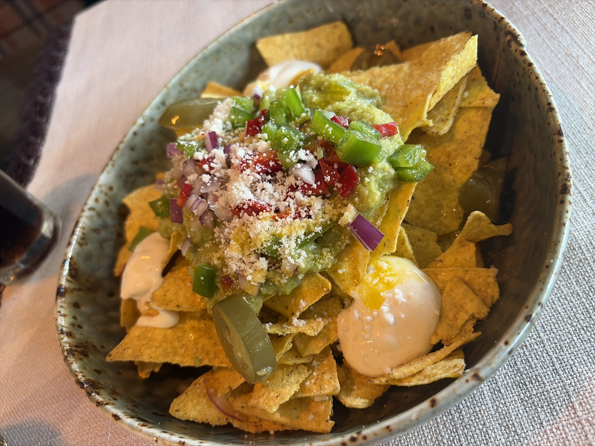 A high-angle, close-up shot depicts a bowl of nachos on a beige textured surface, likely a table. The nachos are arranged in a rustic, gray-toned bowl with speckled detailing along the rim. The contents consist of yellow tortilla chips, topped with a scoop of guacamole, diced red and green peppers, chopped red onion, and a sprinkling of white cheese. A dollop of white sauce is also visible. There are pickled jalapeños scattered around the top layer. A drinking glass is seen blurred in the background. While no location is specified, the description does not indicate Veliko Tarnovo.