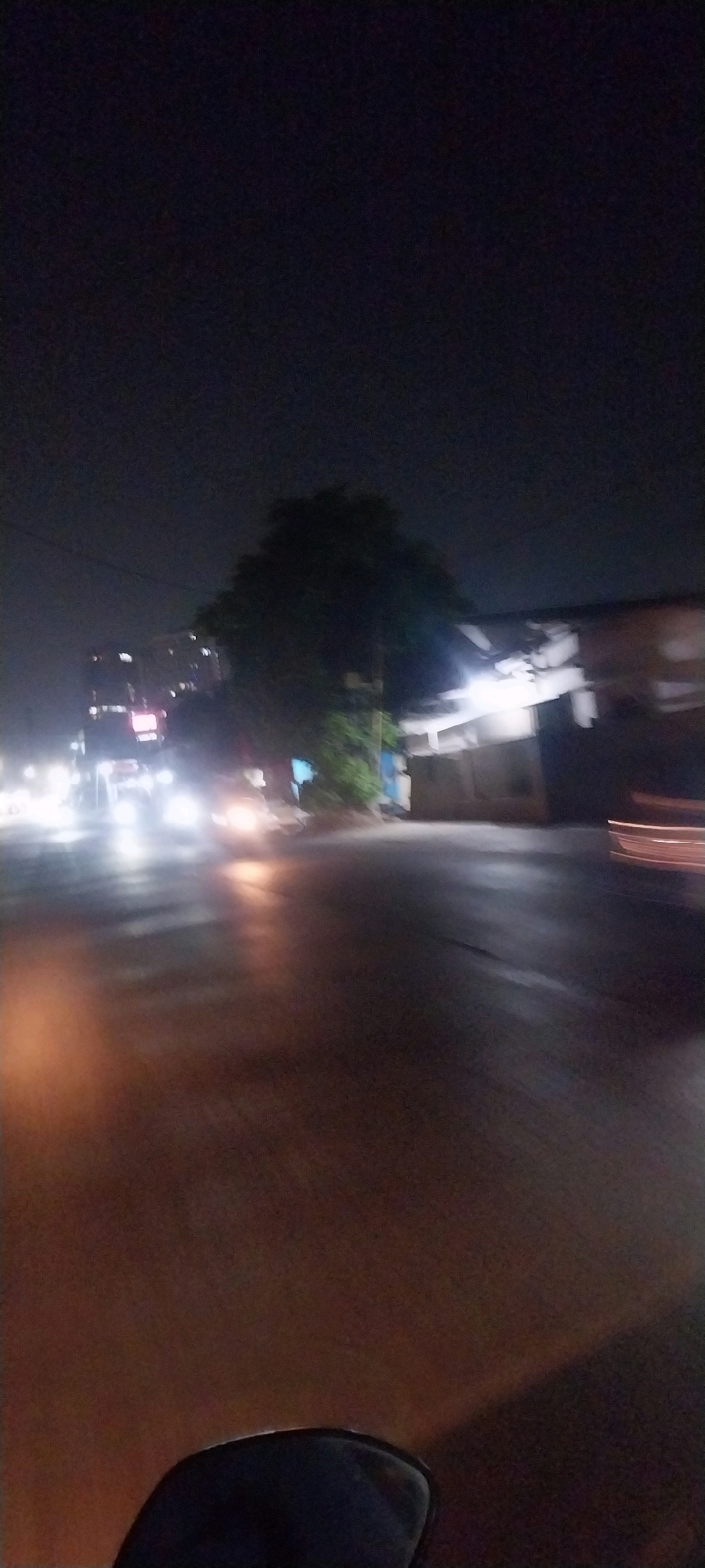 The image depicts a night scene on a wet street in Kelapa Dua, Indonesia. A vehicle's headlights create streaks of light on the road, indicating movement and possibly rain. In the background, a large tree with dark foliage stands near a building with illuminated windows. The overall atmosphere is dark, with artificial lighting from the building and street illuminating parts of the scene. The foreground shows a portion of a vehicle, suggesting the photo was taken from a moving perspective.
