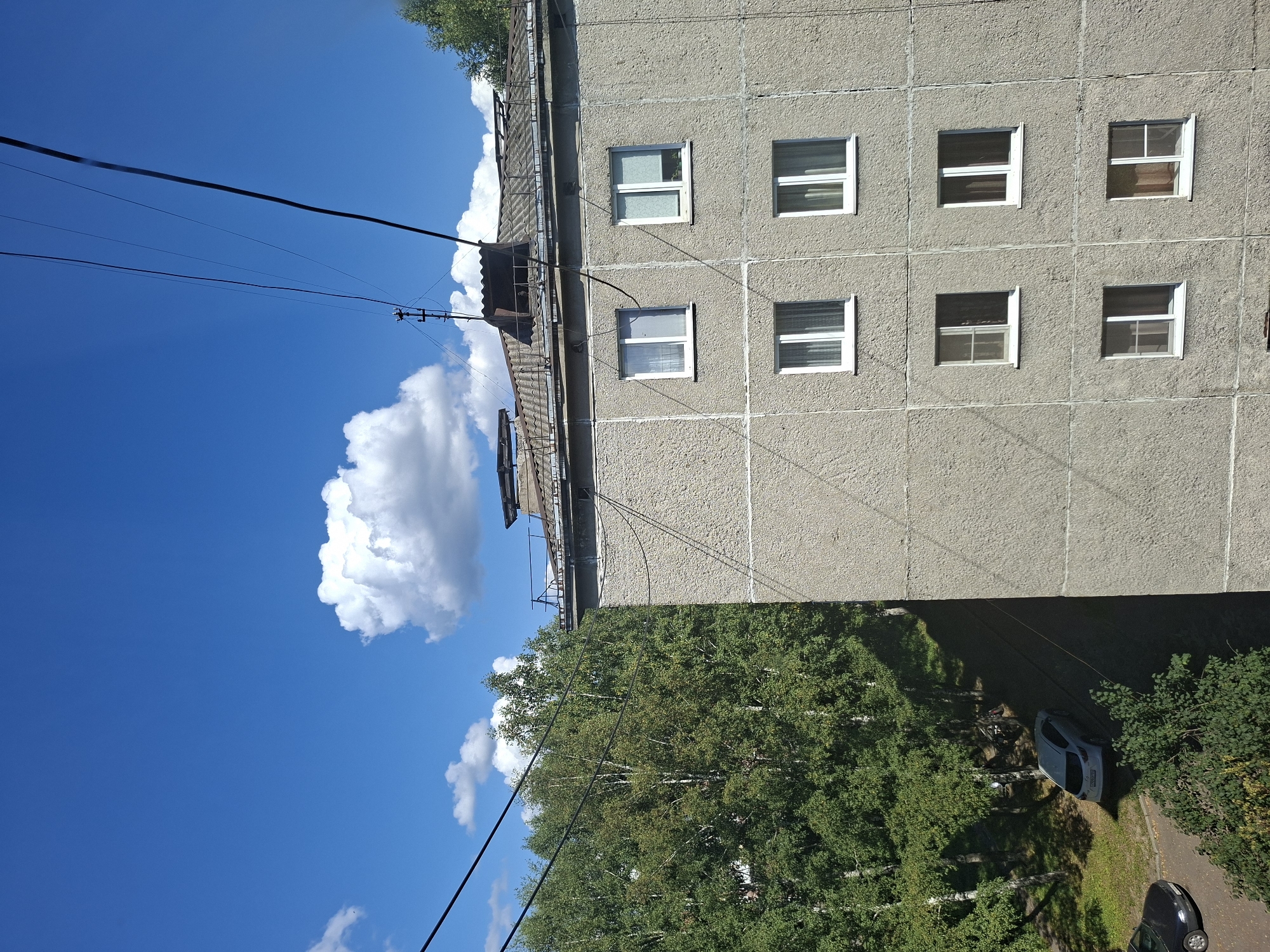 A multi-story building with a rough, gray concrete facade and multiple windows is pictured. The building's roof is visible, covered in gray shingles and some debris. Electrical wires run across the sky, which is a bright blue with scattered white clouds. To the left of the building, a dense cluster of green trees is visible, and below the trees, a white car and a dark car are parked on a paved area. The scene appears to be a residential area in the city of Mezhdurechensk, Russia, on a sunny day.
