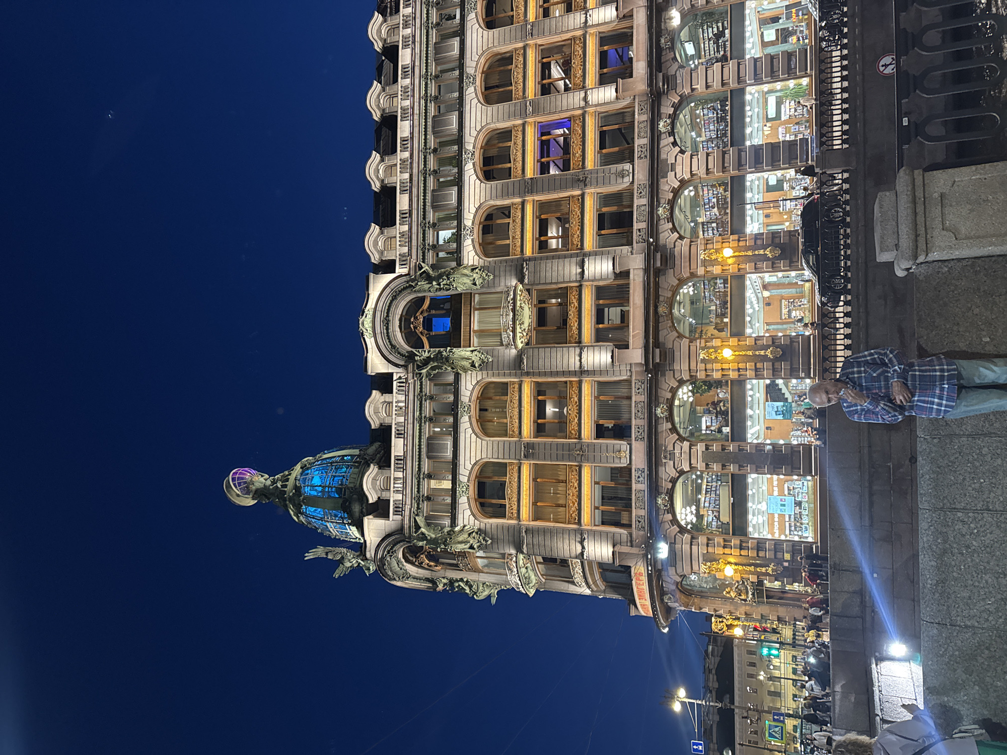 This is a nighttime exterior shot of the Singer House in Saint Petersburg, Russia, a historic building located on Nevsky Prospekt at the crossing with the Griboyedov Canal. The building is illuminated, showcasing its ornate architecture, including arched windows, decorative sculptures, and a prominent glass dome topped with a globe and eagle sculpture, all glowing with blue light. In the foreground, a man in a blue plaid shirt and jeans stands by a stone barrier, holding a cigarette. Behind him, other people are visible in the background, some walking along the street, and others inside the brightly lit ground floor, which appears to be a bookstore. Cars are parked along the street, and streetlights illuminate the scene. The sky is a deep blue, indicating it is late evening or night.