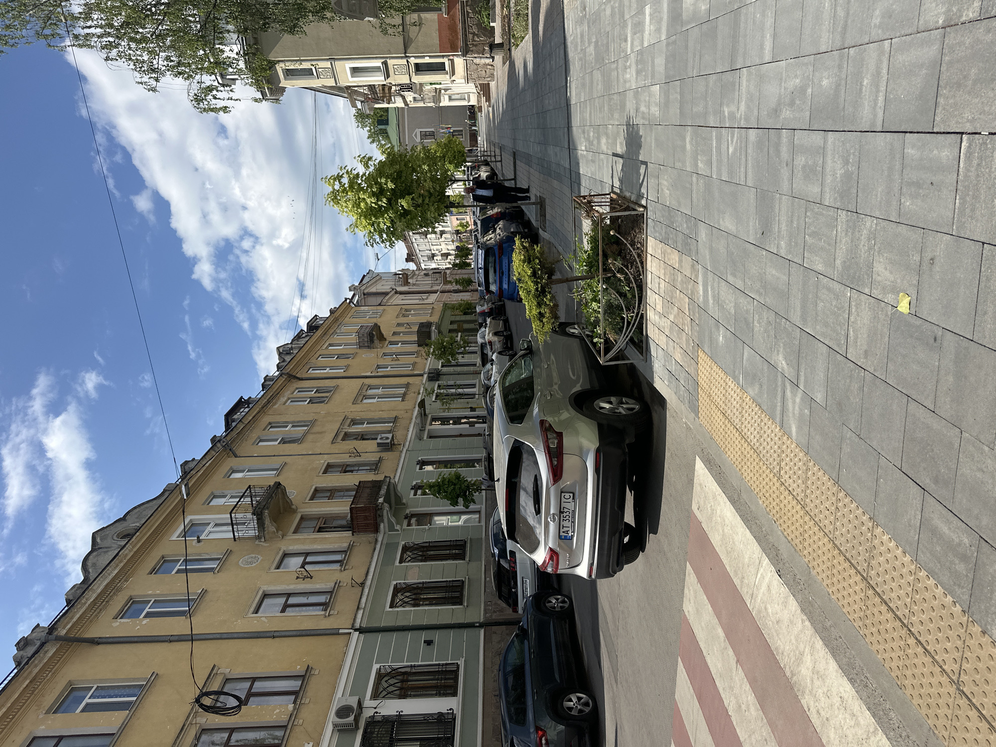The image captures a street in Ternopil, Ukraine. A sidewalk paved with gray rectangular stones is on the right, marked with a crosswalk of alternating red and white stripes. A tactile paving surface for the visually impaired is adjacent to the crosswalk. Cars are parked along the left side of the street, including a silver Nissan Qashqai with a visible license plate "AT 3537 IC." The buildings lining the street are old, multi-story structures with ornate facades in shades of yellow and green. Windows and balconies are visible, some with decorative ironwork. A few trees are planted along the street. A blue sky with scattered clouds is overhead. Some pedestrians are visible in the distance.