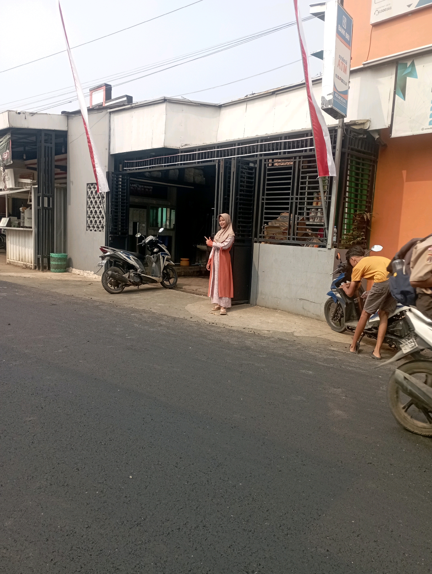 In Pendem, Indonesia, a street scene unfolds in front of a building with a white facade and a gray metal gate. A red and white flag hangs from a pole near the building. To the left, a black and white scooter is parked on the sidewalk. A woman wearing a long, peach-colored dress and a hijab stands near the building, holding an object in her hand. Further to the right, a young person in a yellow t-shirt and gray shorts leans over a blue motorcycle, their back to the viewer. Another motorcycle is partially visible on the right side of the frame, its rear wheel prominent. The street in the foreground is paved with dark asphalt, and the background shows a portion of an orange-colored building.