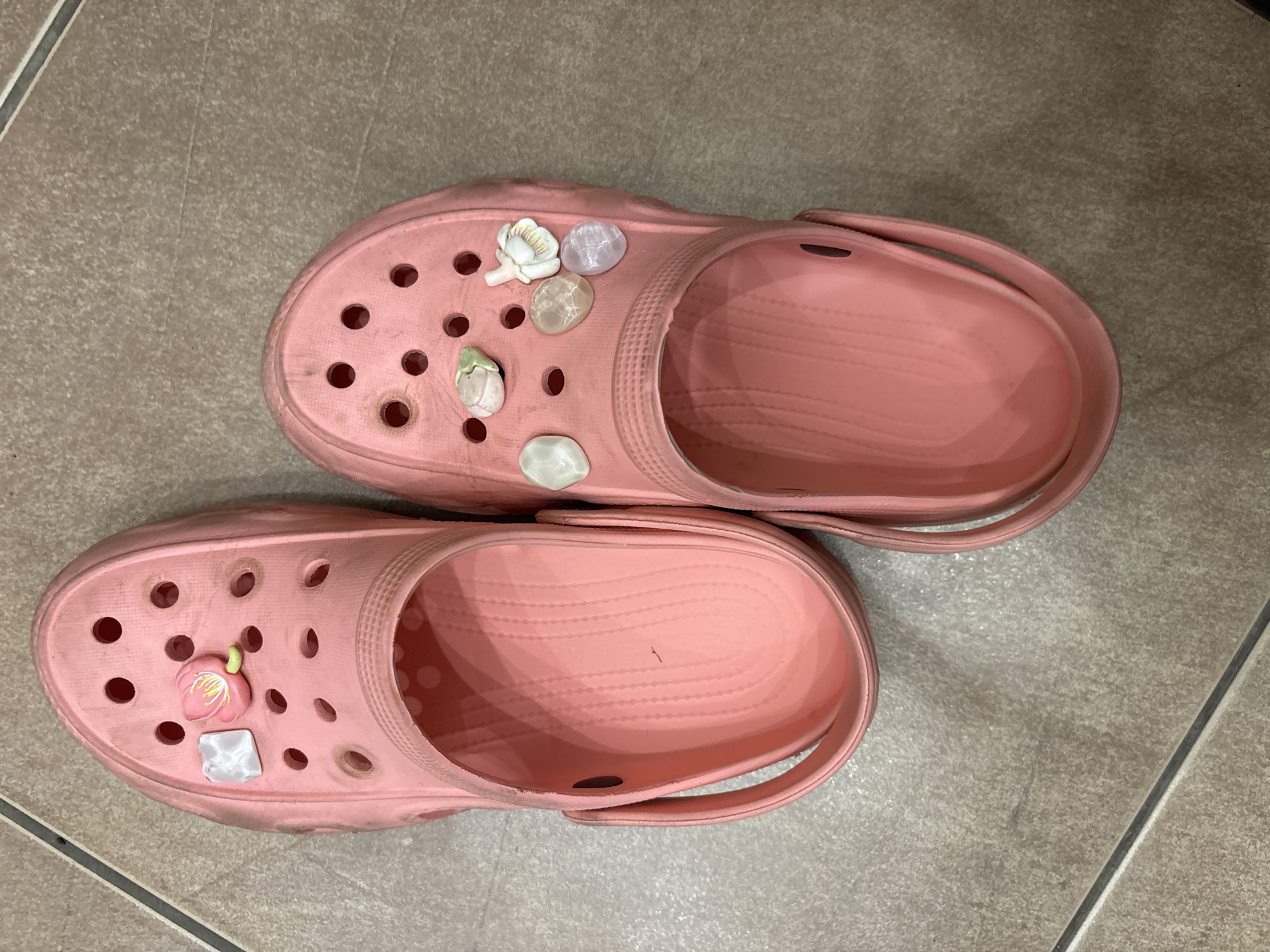 The image shows a pair of pink Crocs on a tiled floor. The Crocs are slightly dirty and adorned with floral and gemstone-like charms. The background is a light brown tile floor, with a grout line visible in the lower right corner.