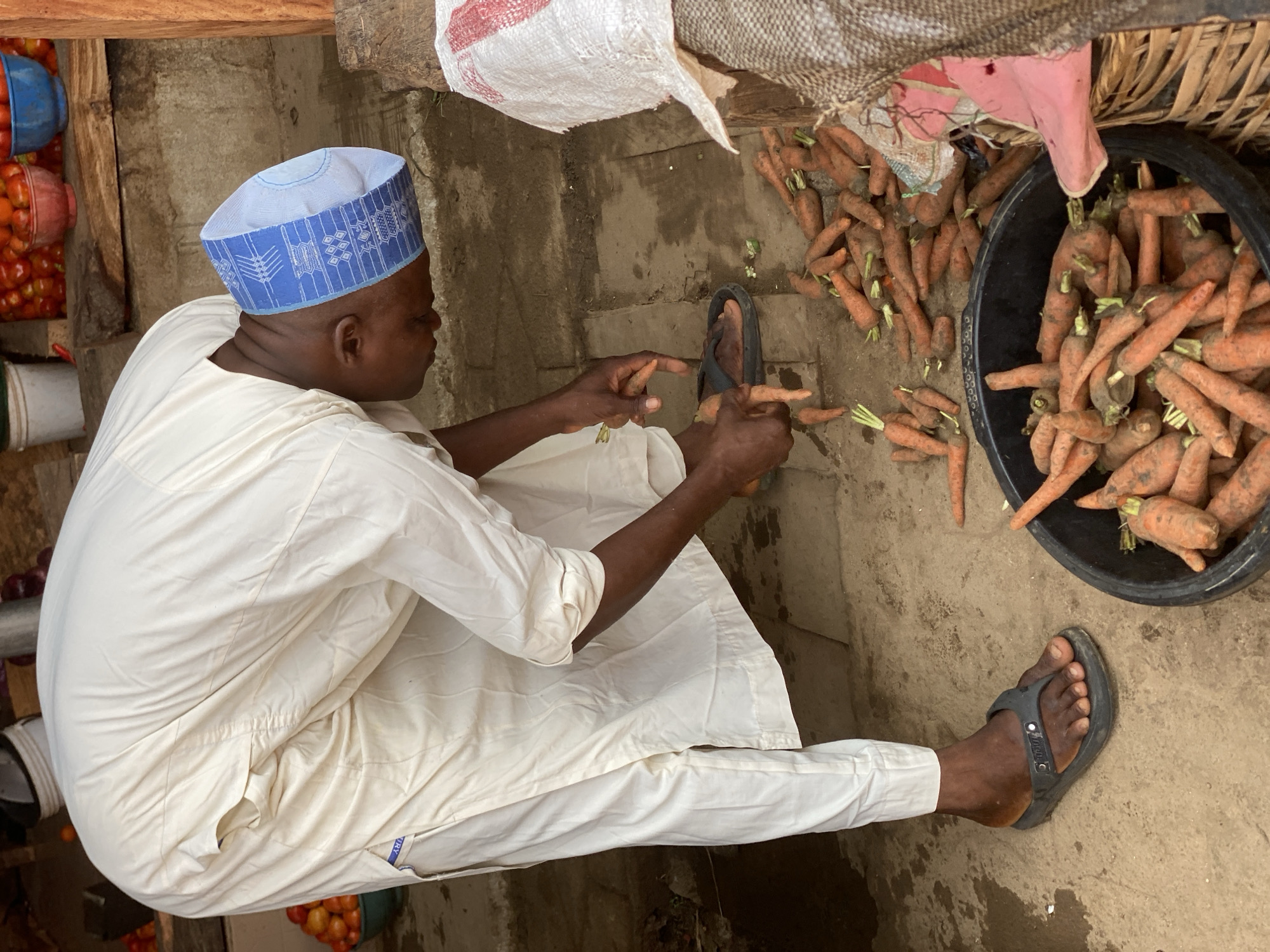 This media file depicts a market scene in Kafanchan B, Nigeria, featuring a person engaged in preparing or sorting carrots. The individual, seen from a high angle and rotated, is sitting on the ground, dressed in a light-colored, possibly white, traditional tunic (agbada) and a blue cap with intricate white patterns.
The person's hands are focused on a pile of small, raw carrots scattered on the dirt or concrete ground. A large quantity of carrots is also contained within a black plastic basin positioned next to them. Many of the carrots retain their green tops, suggesting freshness.
In the background to the left, several colorful plastic containers—blue, red, and white—are visible, filled with numerous red, round fruits, most likely tomatoes. Above the carrots, a white woven sack and a darker, textured bag are present, likely used for produce storage or transport. Wooden beams and posts can be seen, indicating an open-air market stall or a simple structure.
The individual wears black flip-flops, with the word "TOGO" visible on one. A small label with the word "DRY" is visible on the sleeve of the tunic. The scene appears to be a routine activity in a vibrant market setting during daylight hours. The person's expression is not visible as they are looking down at their task.