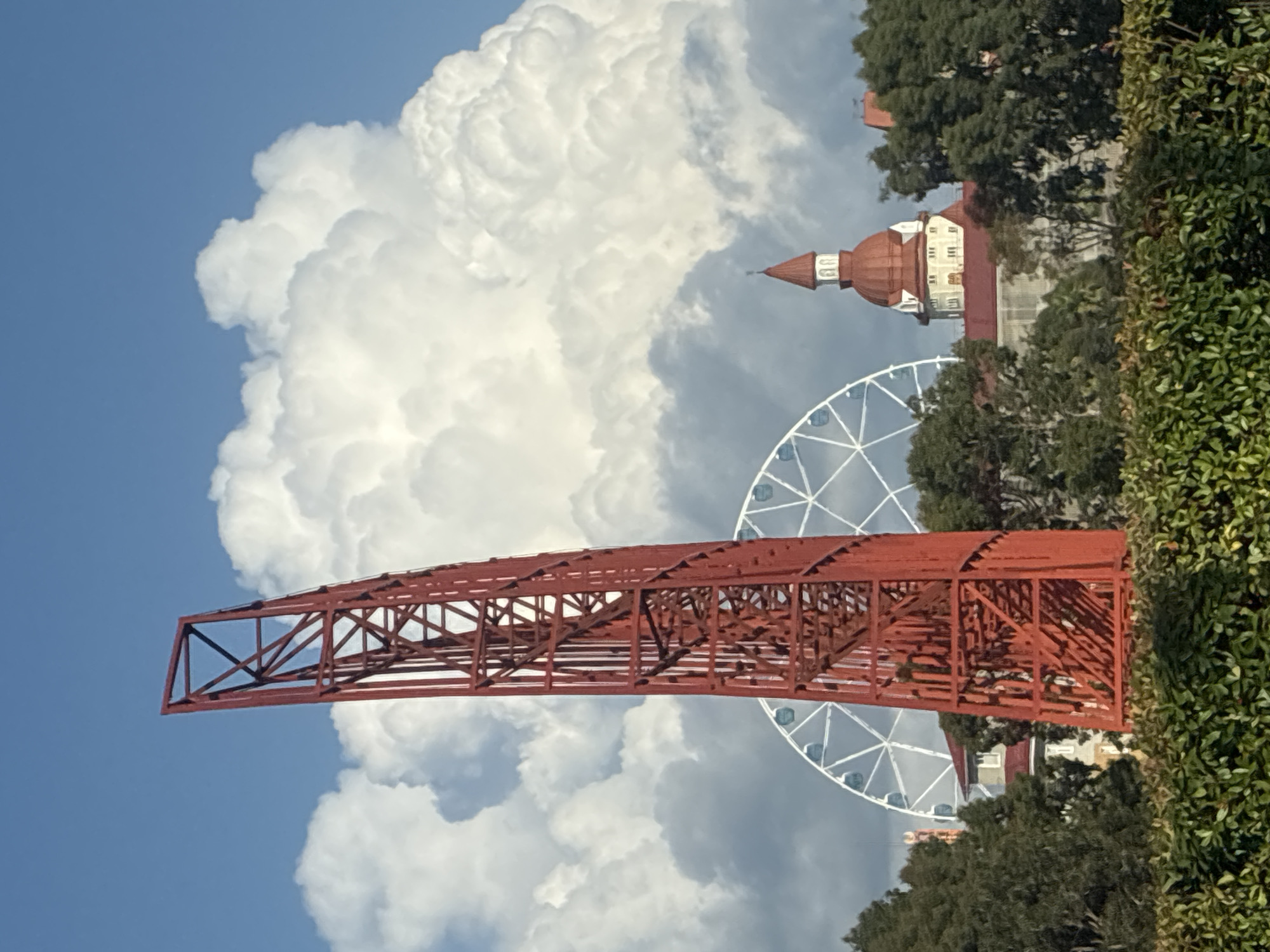 The image depicts a red metal structure in the foreground, resembling a tower or part of a roller coaster, angled slightly to the left. Behind it, a white Ferris wheel is visible, its spokes radiating outwards. Further in the background, a building with a red domed roof and white accents stands out. Lush green foliage forms a hedge in the immediate foreground, obscuring the lower part of the scene. The sky is a clear blue, dotted with large, fluffy white clouds. The scene appears to be an outdoor amusement park or entertainment district. The location is identified as the city of Sirius, Russia.