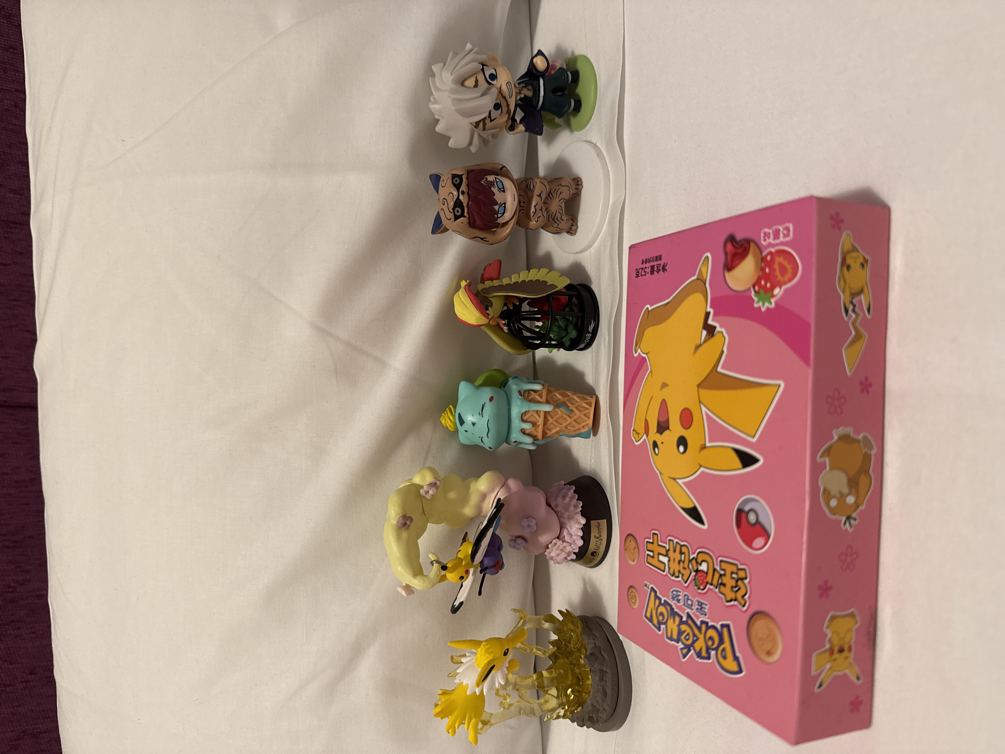 The image depicts a collection of figurines and a box arranged on a white surface, likely a bed or similar flat surface, in an indoor setting.
From top to bottom, the figurines include:
1. A figure with white hair, dressed in a dark suit, on a green base.
2. A figure resembling a character in a hooded cat-like costume with tan-colored skin.
3. A figure appearing to be a bird inside a black cage.
4. A figure with a mint-colored head and brown ice cream cone body.
5. A figure with yellow hair and a figure with insect wings attached to the neck of a figure.
6. A figure with yellow appendages rising from a gray base.
Next to the figures is a pink box featuring Pikachu imagery. The box also includes symbols with a cookie, strawberry, and pokeball. Chinese characters are on the box.
The location in Madrid, Spain, cannot be determined from the provided visual information.