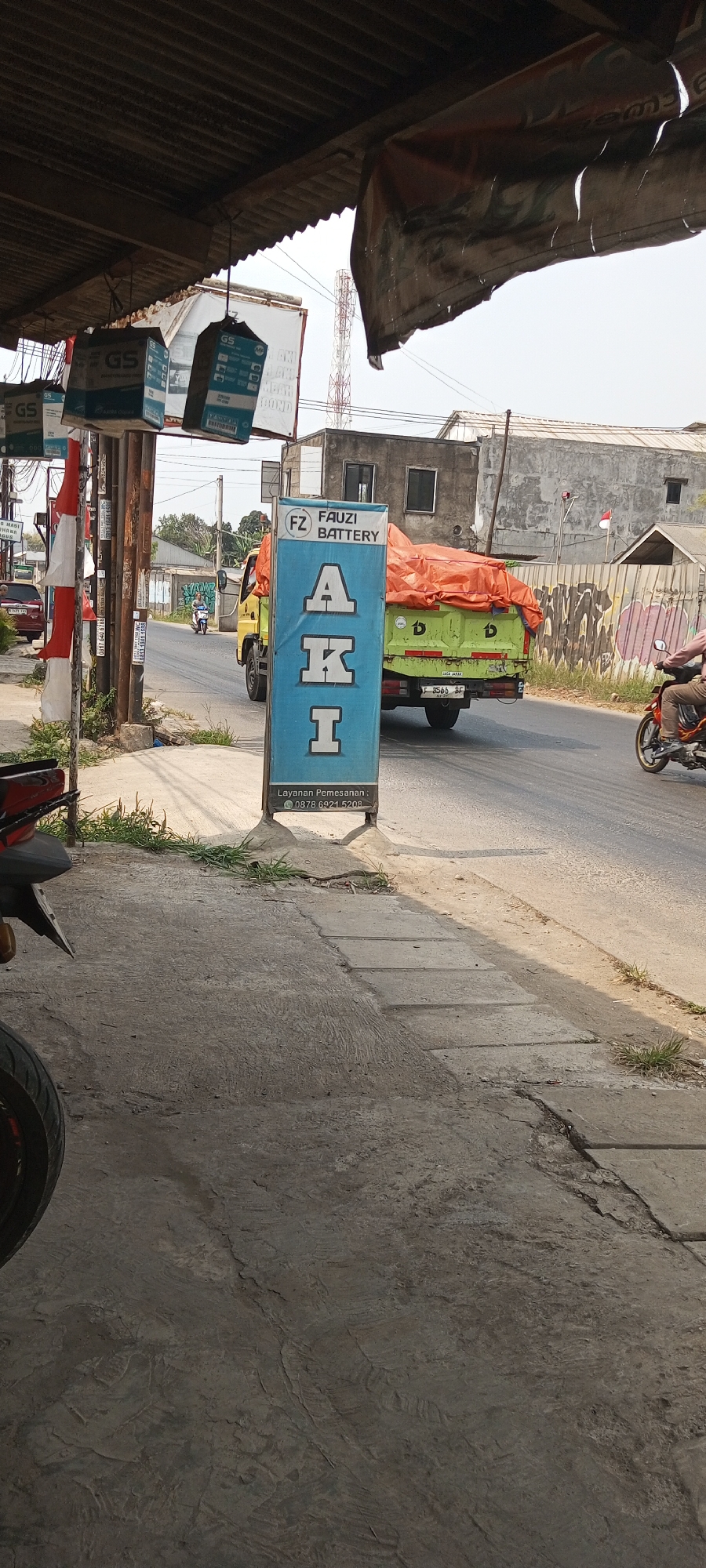 In Setu, Indonesia, a blue sign advertising "FZ FAUZI BATTERY" stands near a road. The sign features the letters "A", "K", and "I" vertically arranged. Below this, "Layanan Pemesanan" (Ordering Service) is followed by the phone number "0878 6921 5208". A green truck with an orange tarp-covered load is moving on the road, its license plate reading "F 2566 BF". A motorcycle rider is visible on the right side of the road. In the background, buildings and trees are present, with a communication tower in the distance. Several "GS" branded battery boxes are suspended from an overhead structure on the left side of the image. The foreground shows a concrete sidewalk.