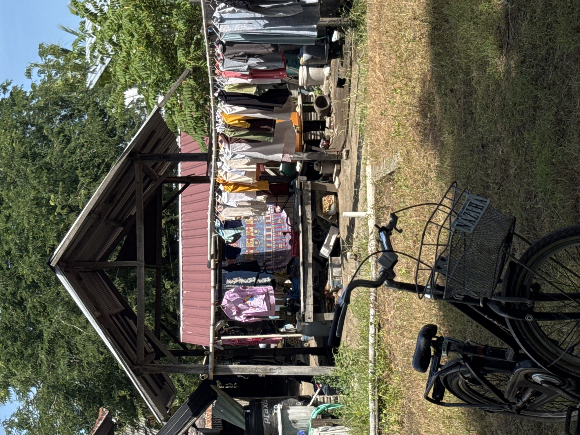 The image shows a bicycle parked on grass in what appears to be a yard. Behind the bicycle, there is a wooden structure covered with a red-colored roof, with clothes hanging from it. The surrounding environment has trees and buildings. The location is in Unknown, Indonesia.