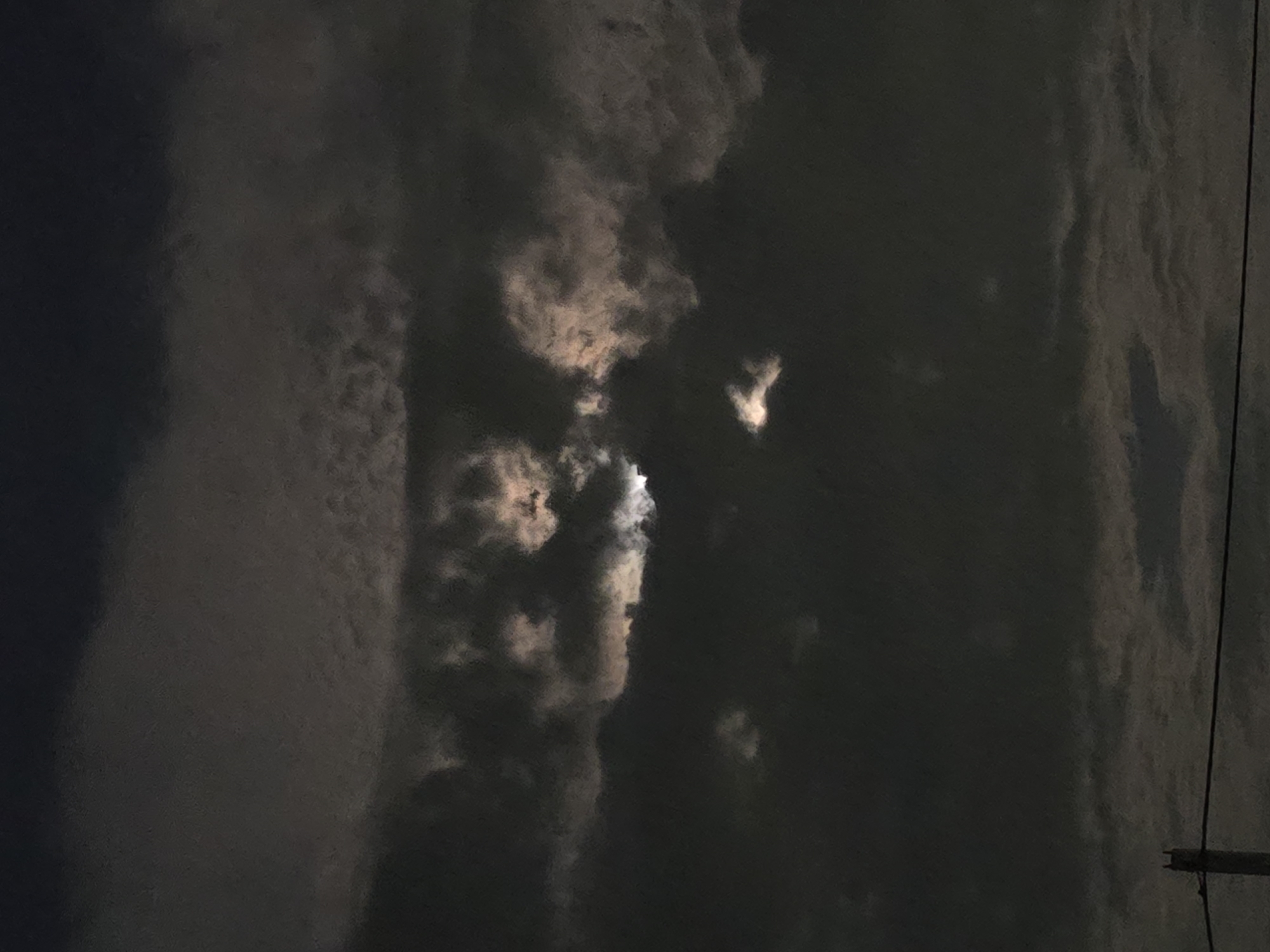 The image shows a night sky with a moon partially obscured by dark, cloudy layers. The moon's light filters through gaps in the clouds, creating luminous patches that highlight the texture of the overcast sky. Below the clouds, the silhouette of a telephone pole and a wire are visible against the faint glow of the horizon, suggesting an urban or semi-urban environment. The scene evokes a sense of mystery and a potentially melancholic atmosphere, typical of a cloudy night. The location provided, Ермолаевский Затон, Russia, places this scene in a specific geographical context, suggesting it could be a residential area or a more rural setting on the outskirts of a town or city in Russia.