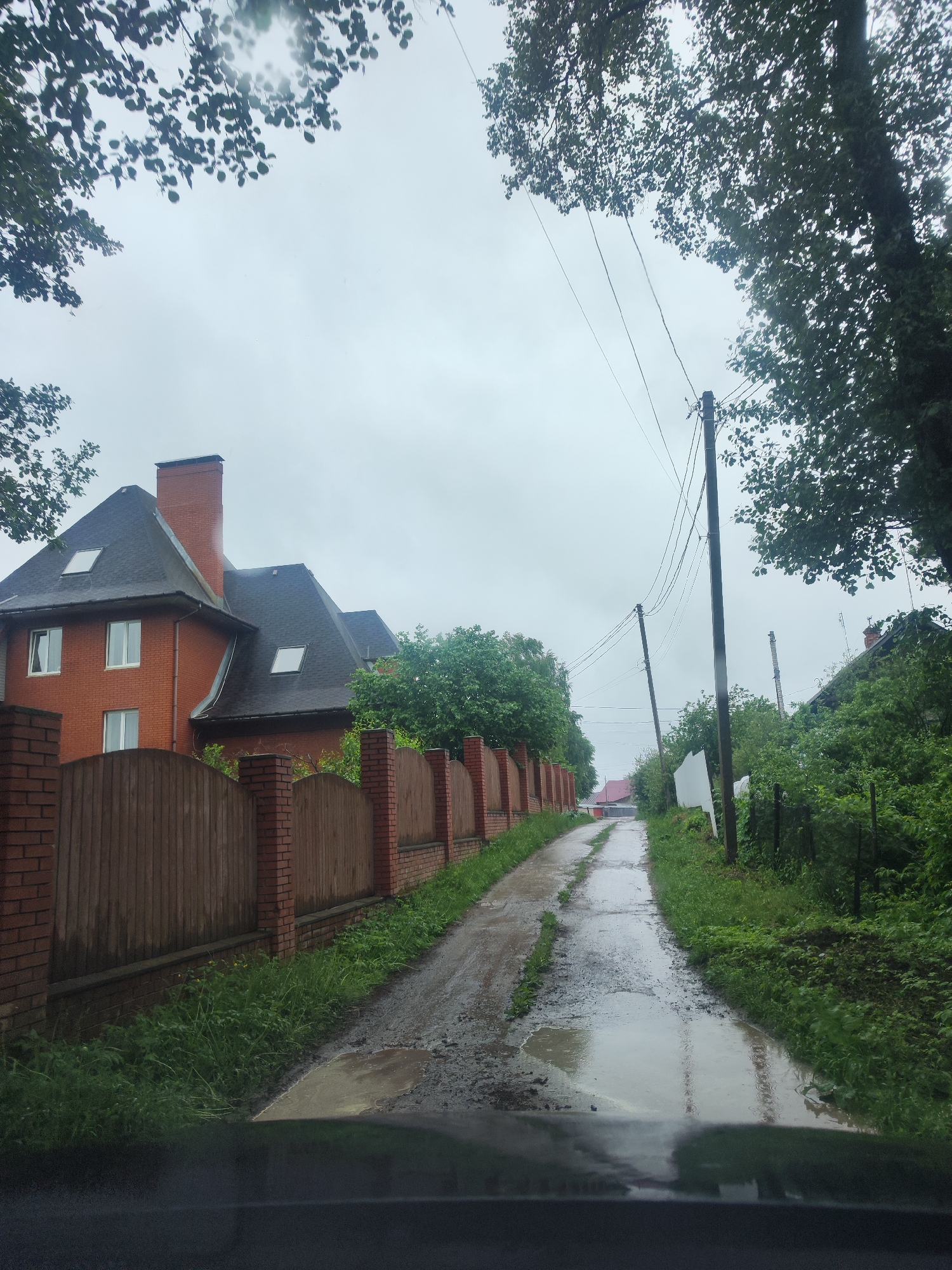 A wet, narrow road stretches into the distance, bordered by a brick and wood fence on the left and dense vegetation on the right. A large brick house with a dark roof stands behind the fence on the left. Utility poles line the right side of the road. The sky is overcast and foliage is visible overhead. The scene appears to be a residential area.