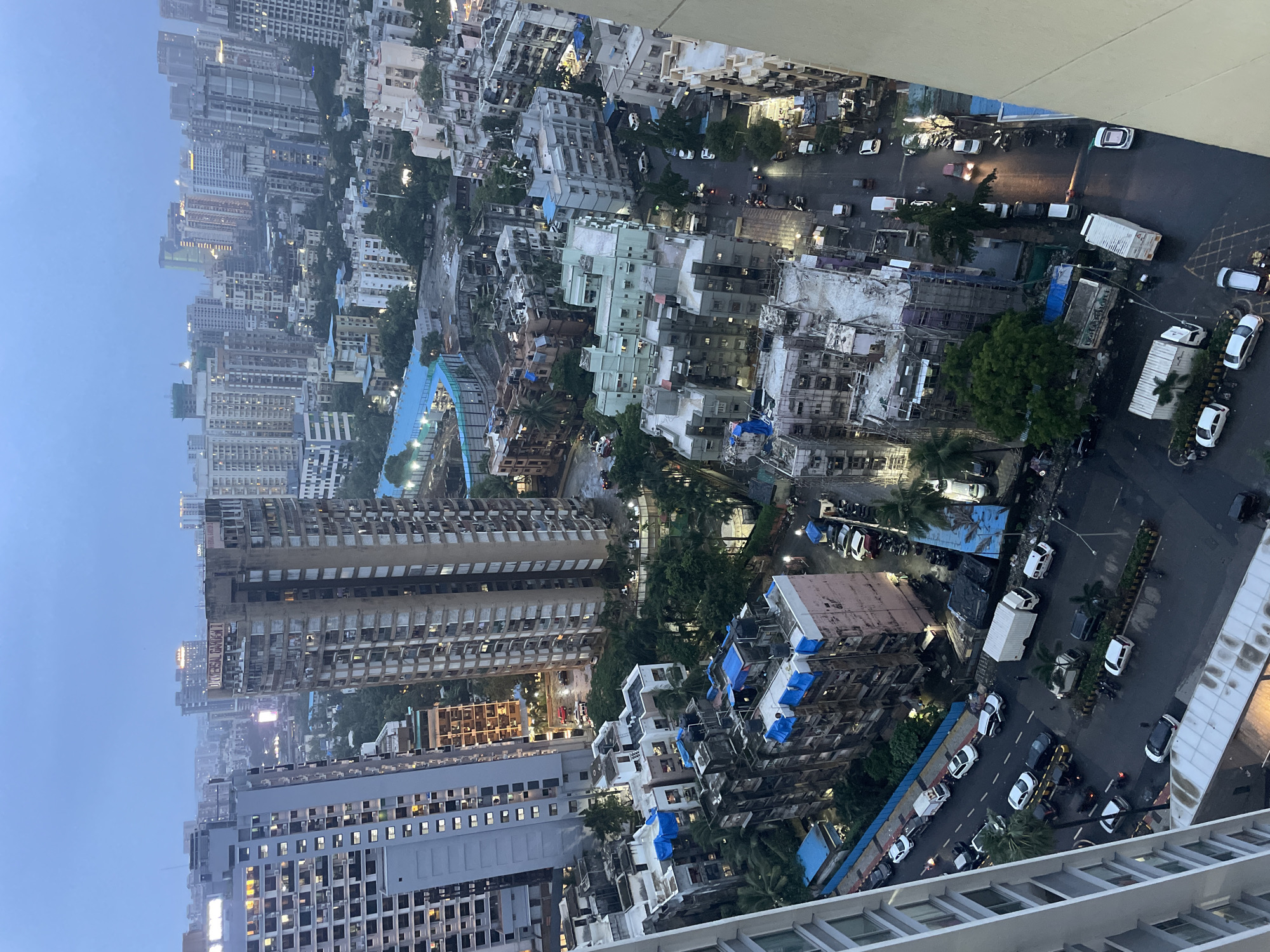 This is an aerial, high-angle photograph of a dense urban environment in Mumbai, India, likely during or after a rain shower. The sky is overcast and a dull gray, suggesting a gloomy day. The scene is dominated by numerous tall residential buildings, varying in height and architectural style, packed closely together. Some buildings exhibit signs of age and wear, with some showing colorful tarps on their roofs.
Below the towering structures, a street is visible, wet and reflecting the lights from the buildings and vehicles. Several cars, trucks, and motorbikes are navigating the street, some of which appear to be parked along the side. The street is lined with more buildings, some appearing to be older, lower-rise structures, and others continuing the theme of high-rise apartments. Greenery, primarily in the form of trees, is interspersed throughout the urban landscape, offering a contrast to the concrete and glass. Construction scaffolding is visible on at least one building. The overall impression is one of a bustling, densely populated city, characterized by its verticality and the constant presence of vehicles.