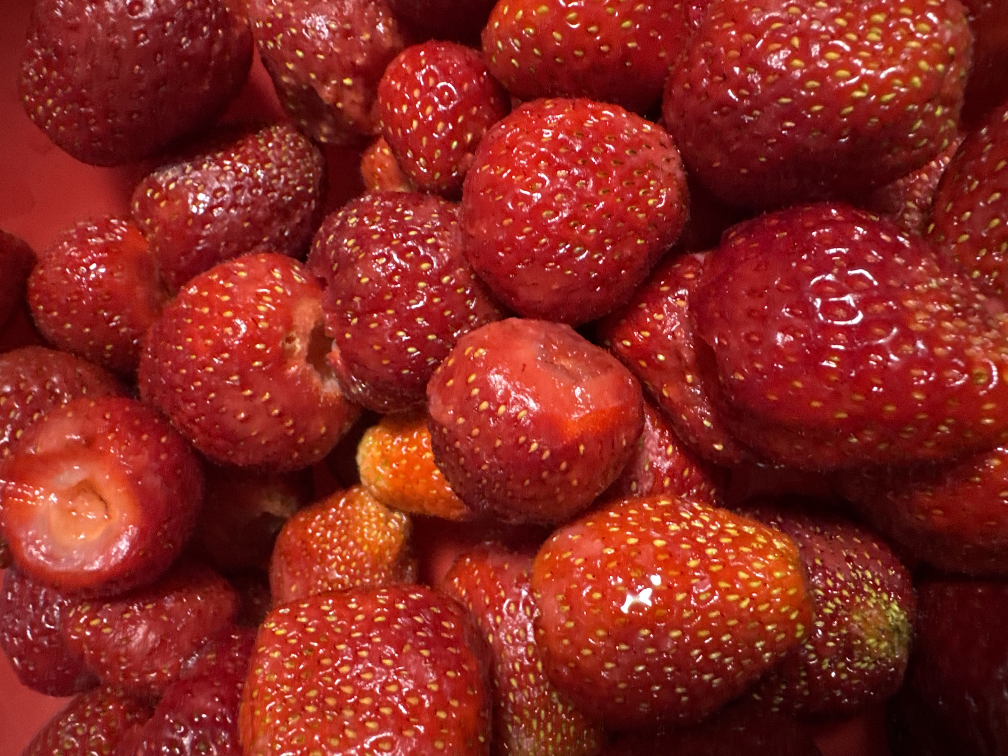 This is a close-up, overhead shot of a pile of fresh strawberries. The strawberries are a vibrant red color with visible seeds. Some are whole, while others appear to be cut in half, revealing their white interior. The image is likely taken indoors, possibly in a kitchen or a food market. The background is a blurred red surface, consistent with a bowl or container for the fruit. There is no indication of any specific location within the city of Chaikovsky, Russia, or any people present in the image.