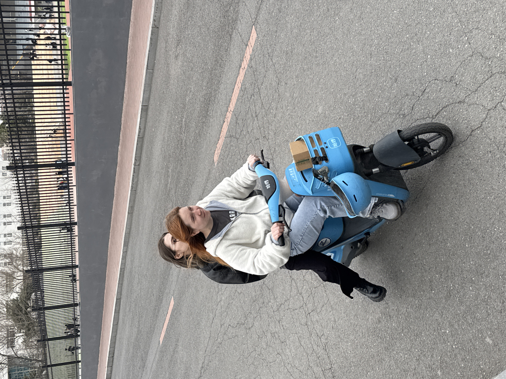 Two young women are riding a blue electric scooter on a paved road. The person in front is wearing a white hooded jacket and jeans and holding the handlebars. The person behind is wearing a black jacket and black pants. A building and a fence are to the left of the road. The scooter has a blue basket on the back.
The location in the city of Zhongtun, China, is not verifiable from the image.
